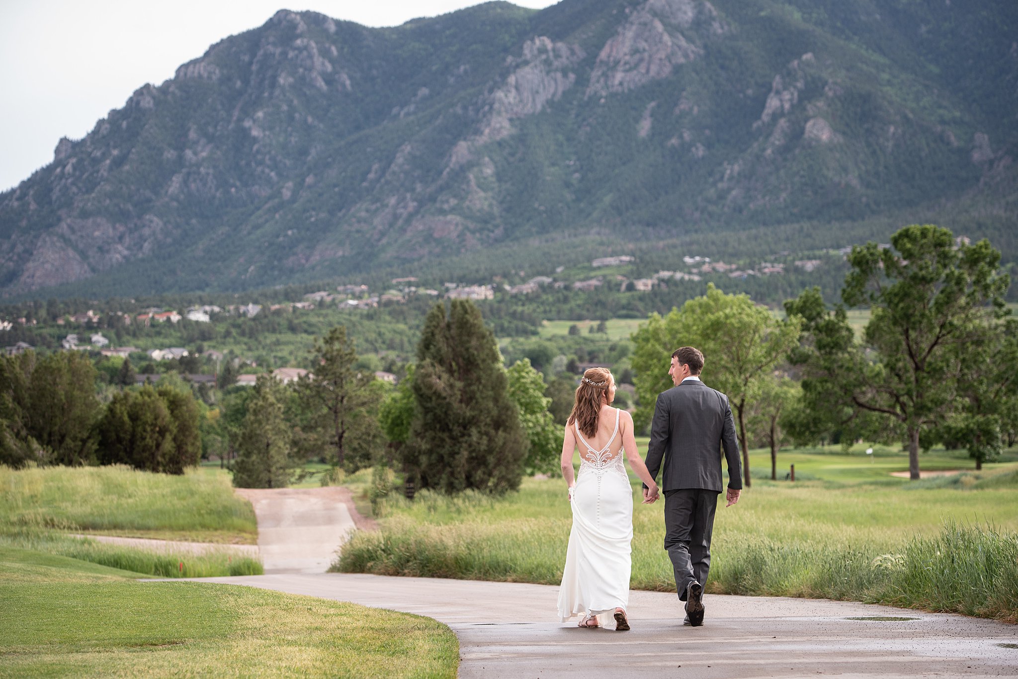 Bride and groom walk down a concrete path through a fields of grass with a mountain backdrop Cheyenne Mountain Resort