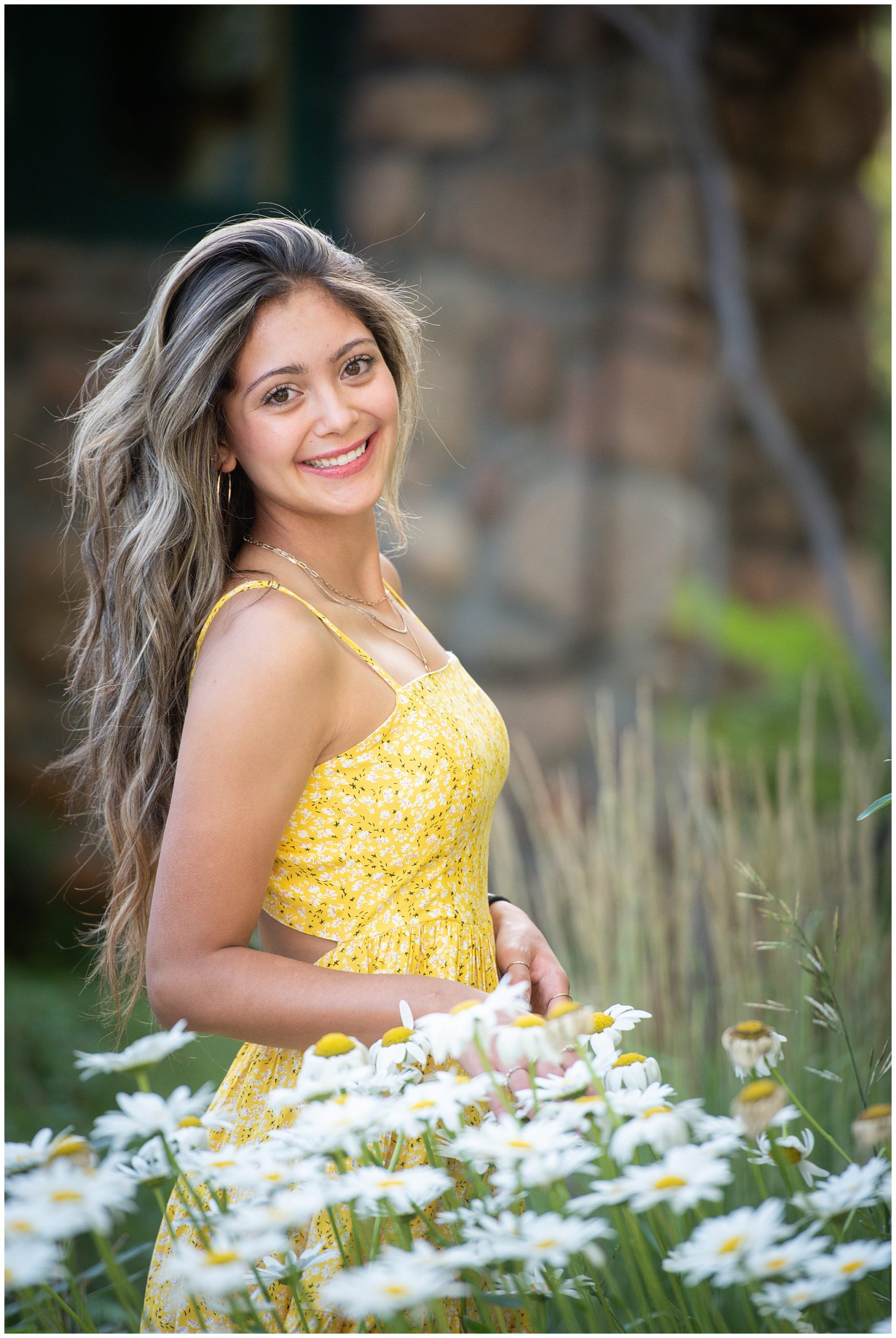 A high school senior wears a spaghetti strap yellow dress in a field of white wildflowers