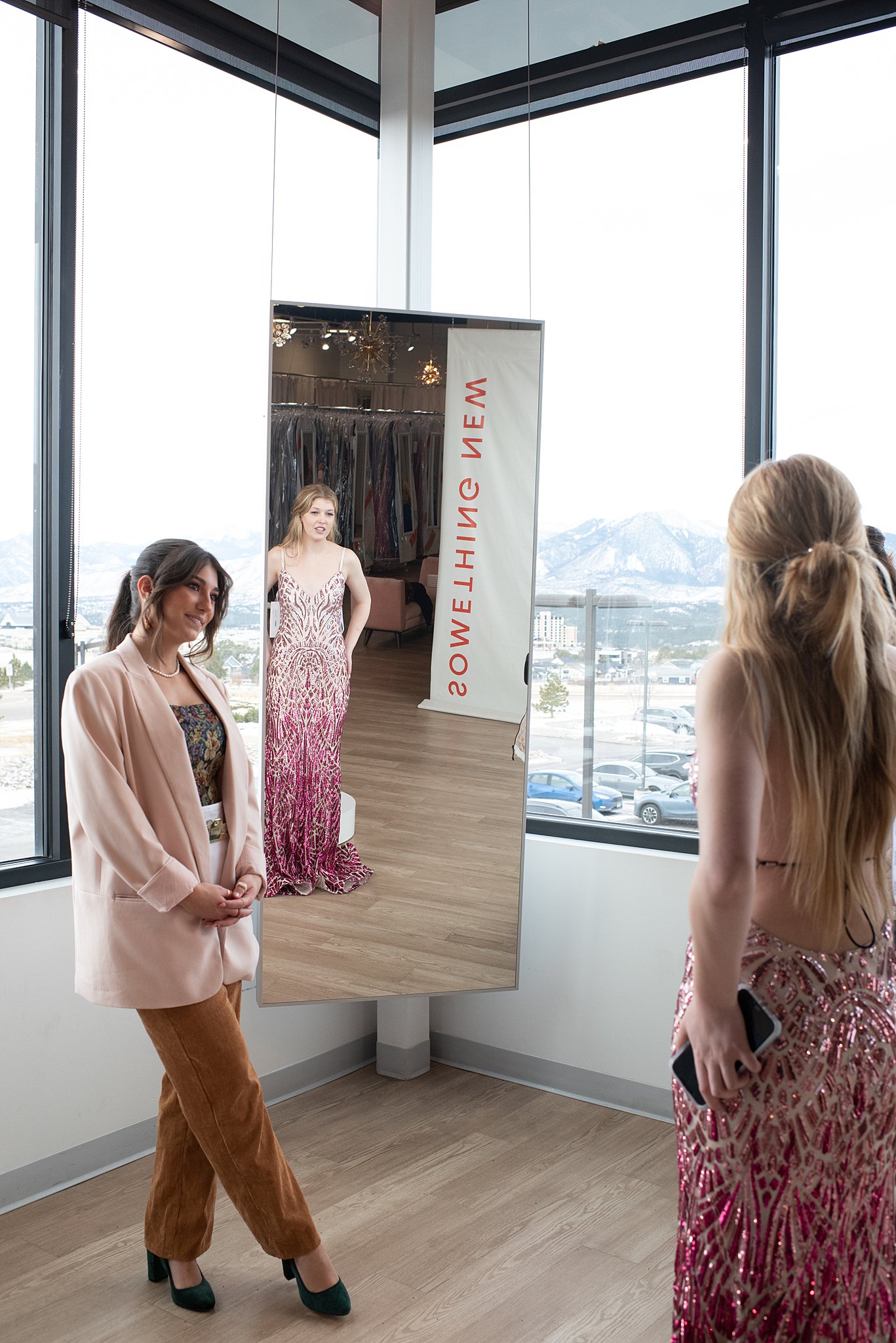 A woman in a pink jacket looks on as a woman tries on a pink and white backless gown Something New Boutique