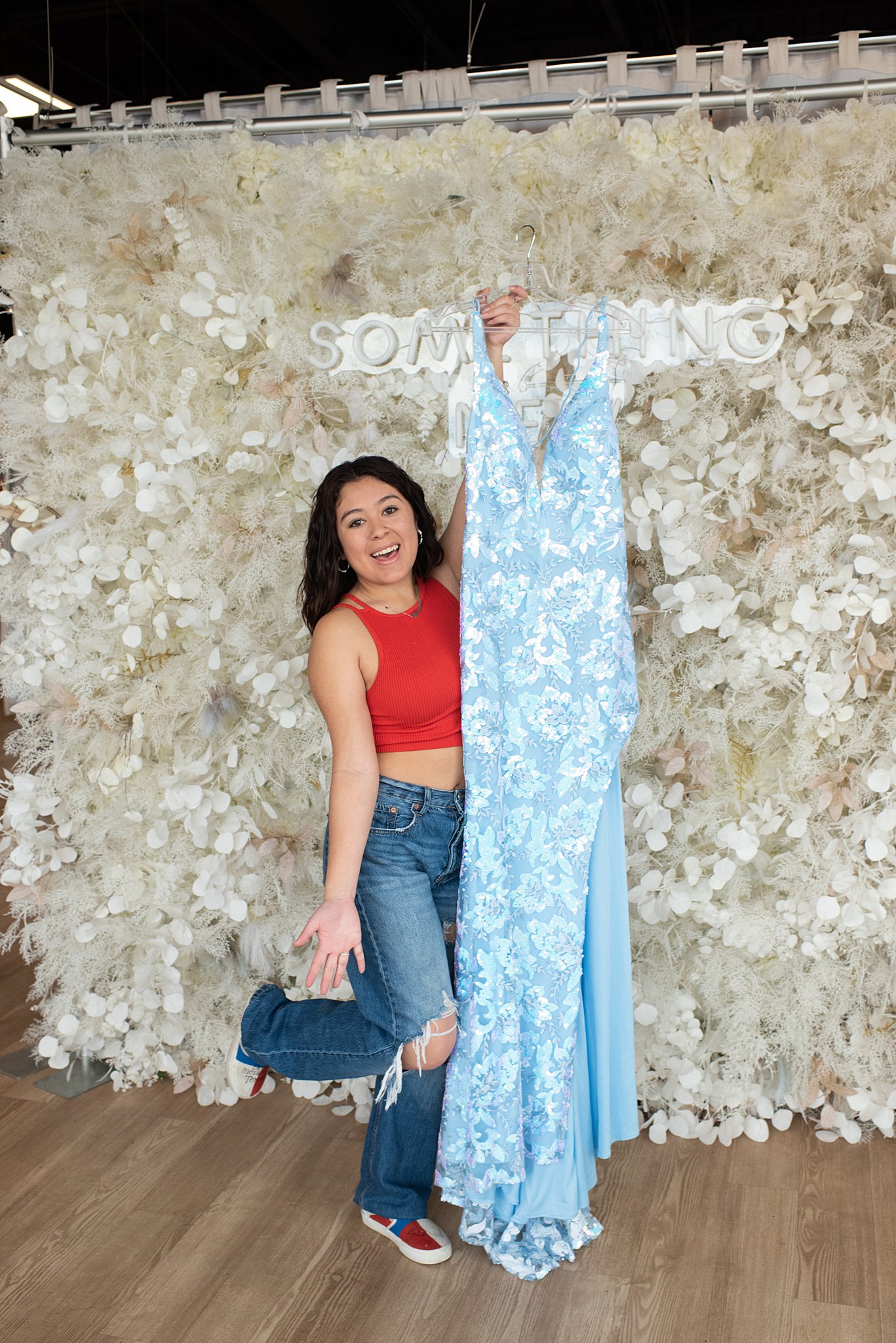 a woman in jeans stands in front of a floral backdrop holding her new blue floral gown