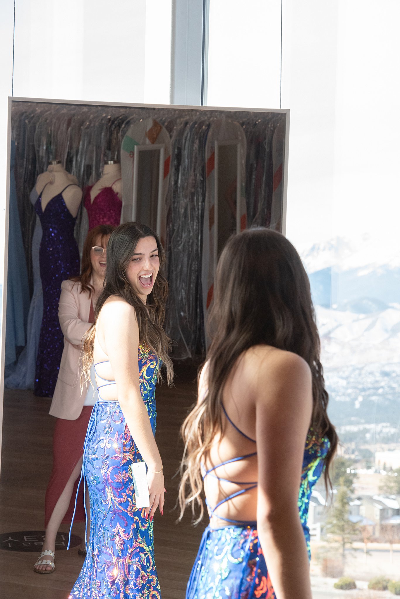girl laughs and enjoys trying on a backless blue gown in front of a mirror Something New Boutique