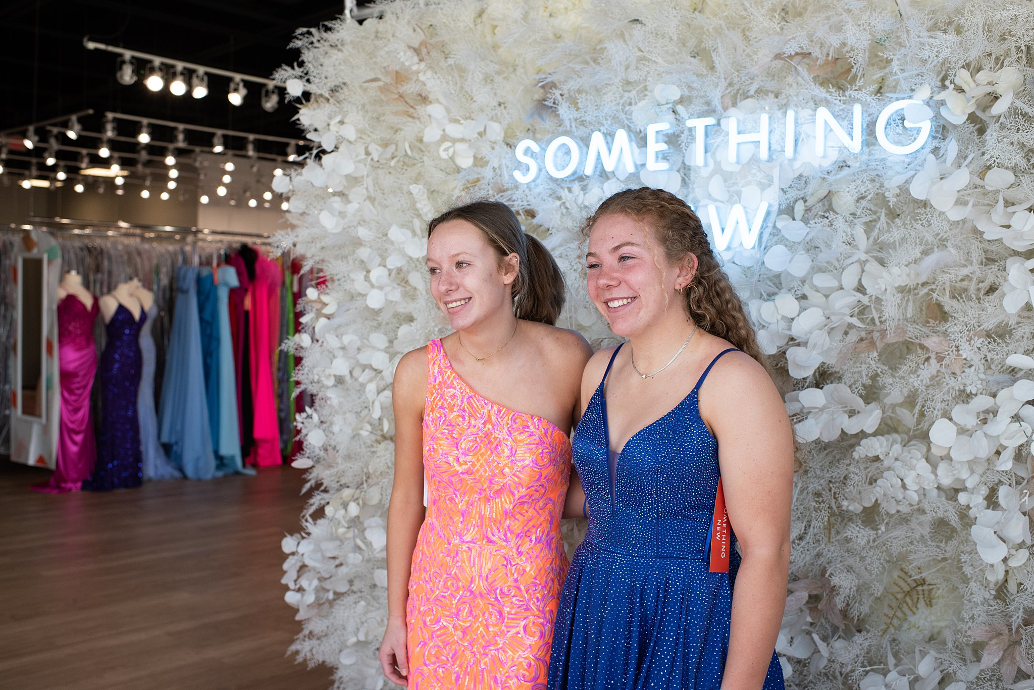 friends stand in front of a floral wall wearing their new gowns Something New Boutique