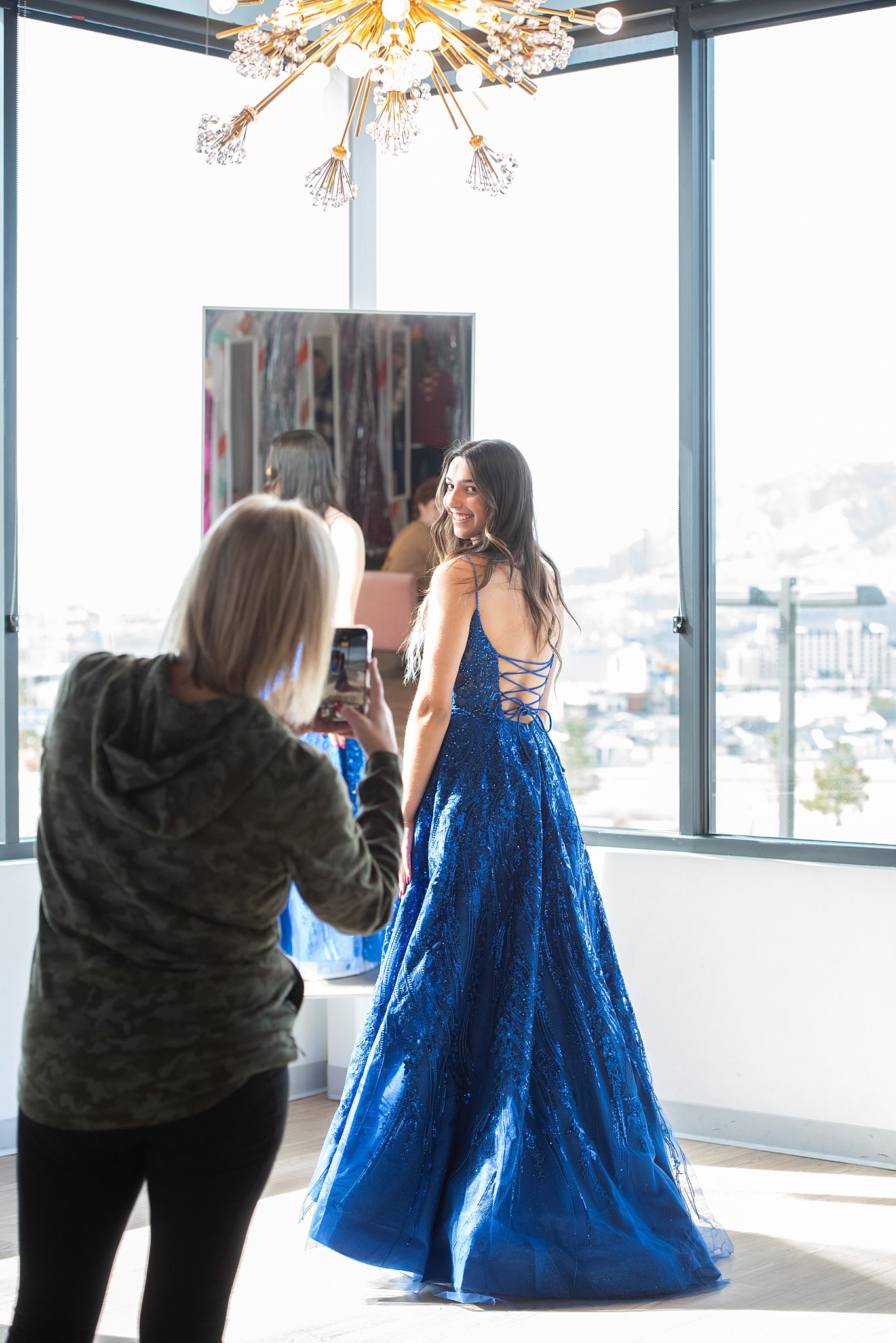 Woman in blue gown stands in front of a mirror while mom takes her photo with a phone