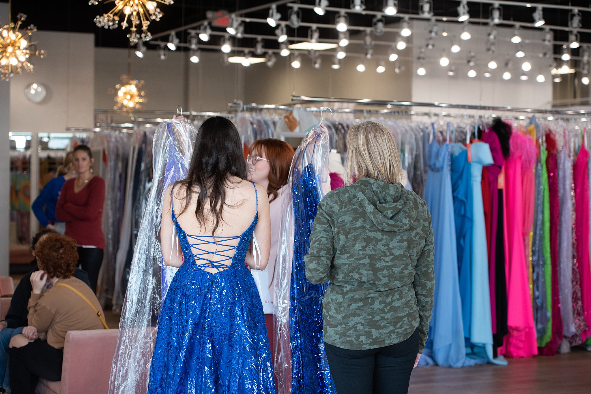 Woman with dark hair tries on a blue gown while being shown other options to choose from Something New Boutique