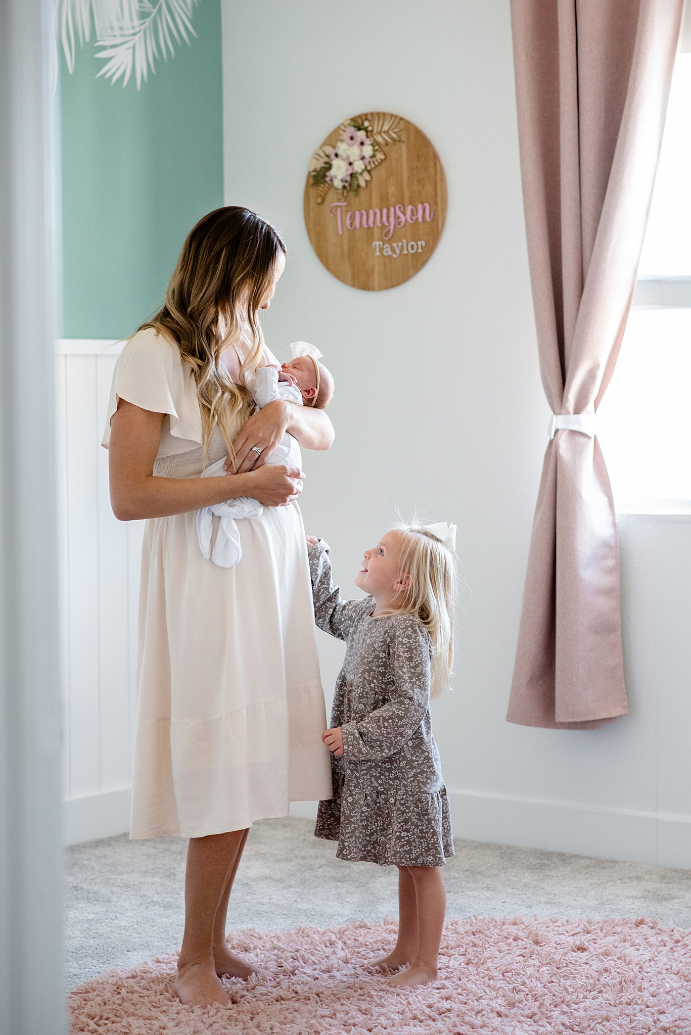 A mother in a cream dress stands in a nursery holding her newborn baby while her toddler daughter stands with her thanks to beautiful births