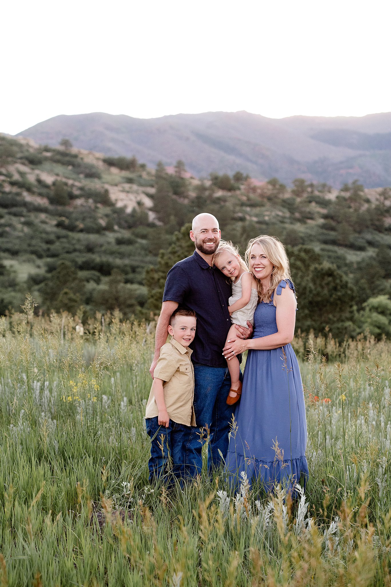 A mom and dad stand in a pasture in the mountains with their toddler daughter and son after visiting club pilates colorado springs