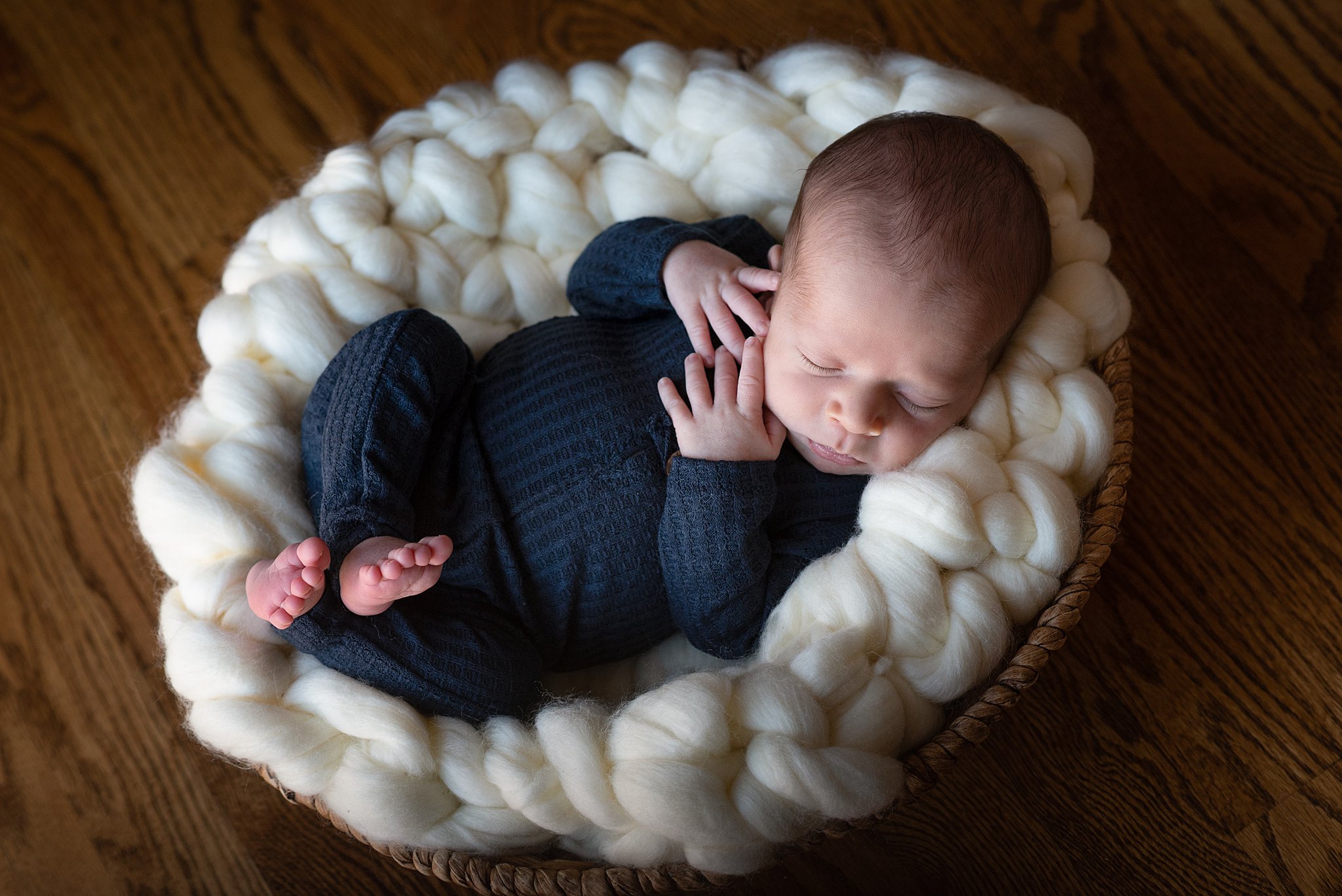 A newborn baby in a blue onesie sleeps in a wicker basket on a wood floor thanks to Colorado Mountain Doulas