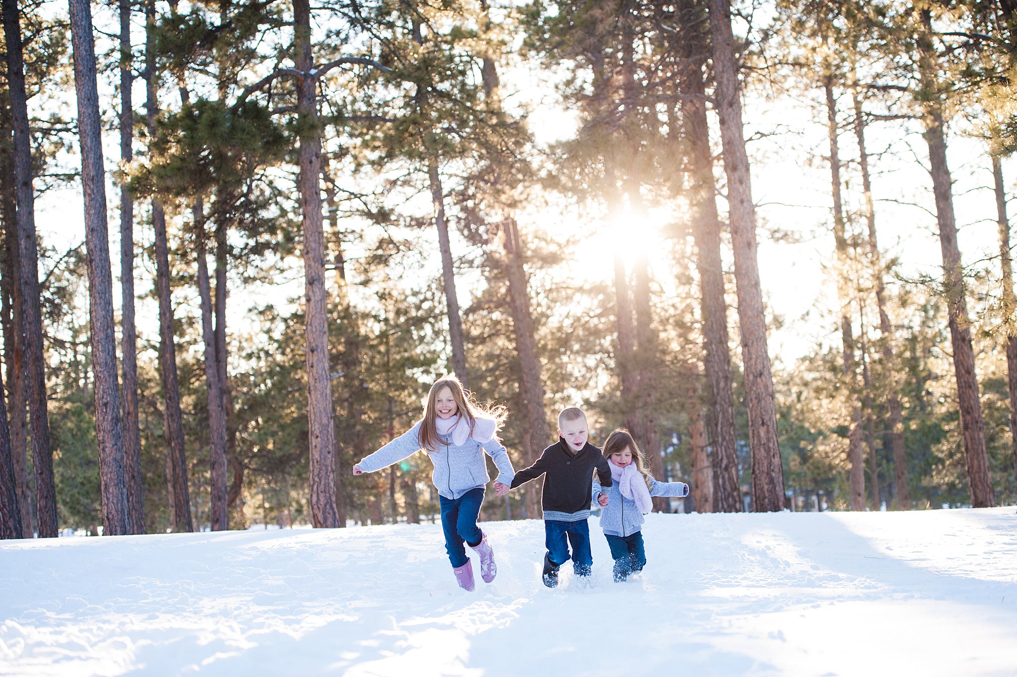 Three young siblings run through the snow in a forest park at sunset during one of the Colorado Springs Family Activities
