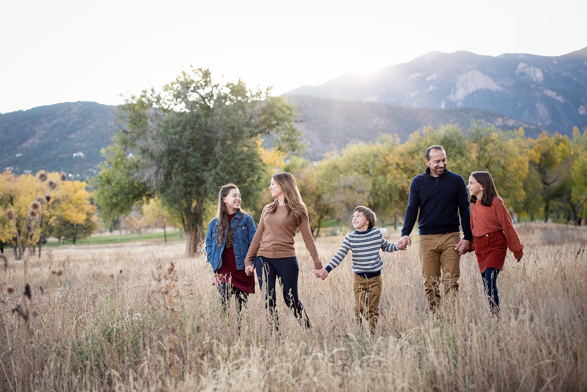 A family of five walk through a field of tall golden grass at sunset while laughing and holding hands at one of the colorado springs family resorts