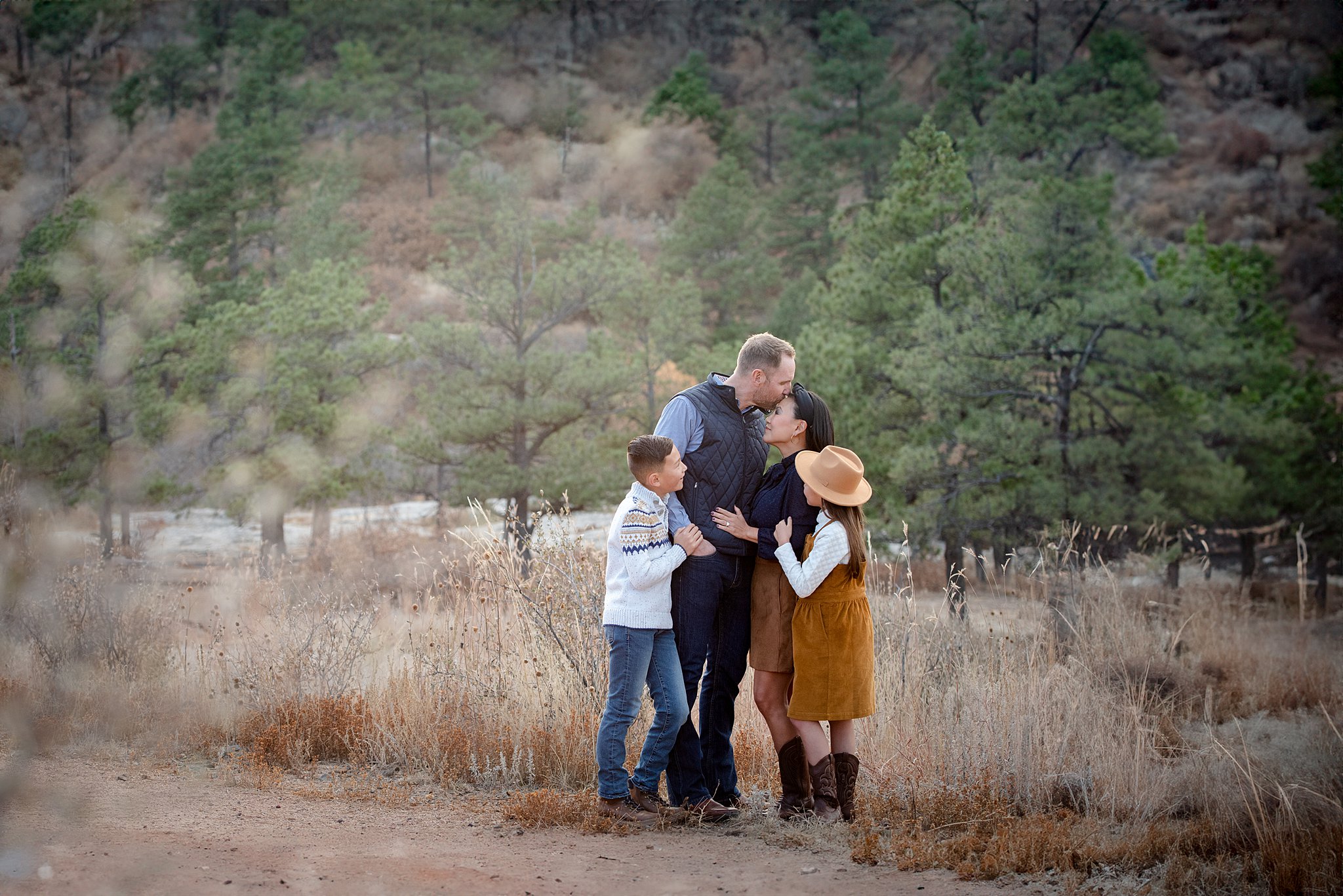 A father kisses the forehead of his wife while their two children hang at their side on a mountain trail after visiting a colorado springs pediatric dentistry