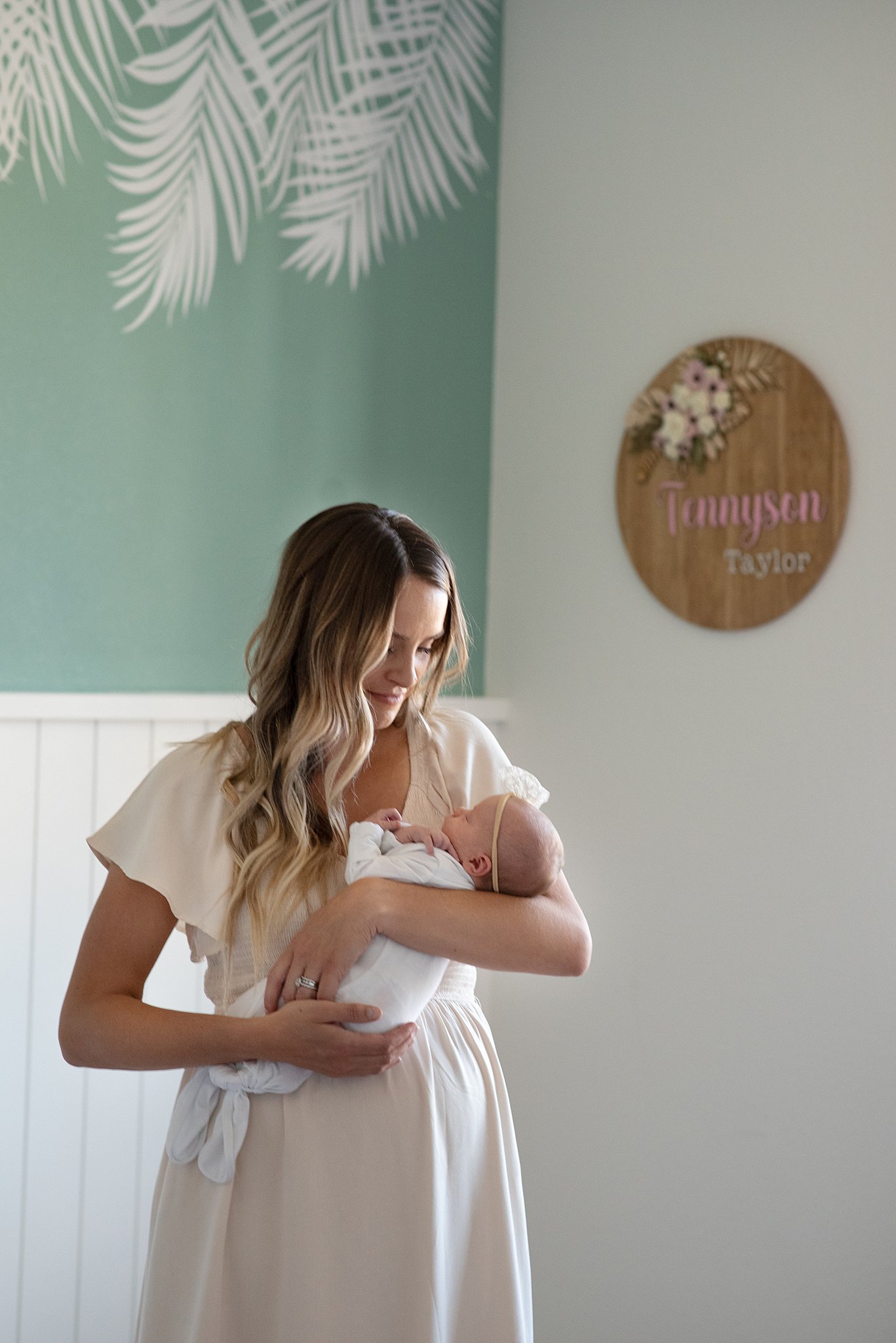 A mother in a cream dress smiles down to her newborn baby while standing in a nursery