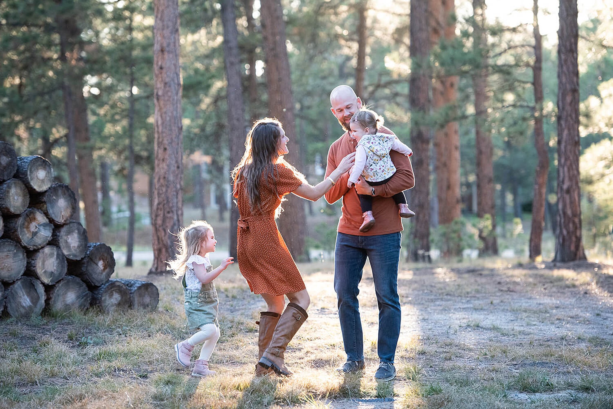 A mom and dad play with their two toddler daughters in a forest at sunset