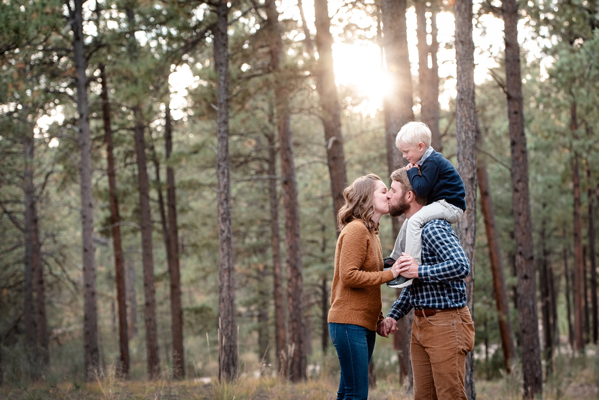 A mom and dad kiss while standing in a forest holding hands with their toddler son on dad's shoulders after some mommy and me classes Colorado Springs
