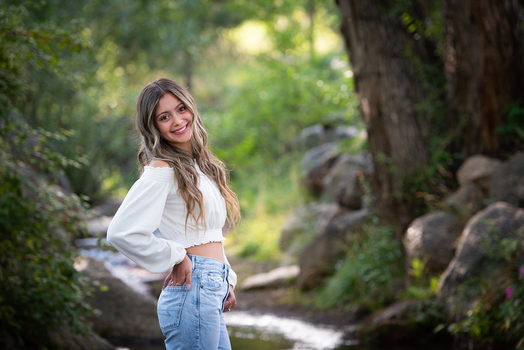 A high school senior rests a hand on her hip while standing by a creek in jeans and white shirt after getting tutoring colorado springs