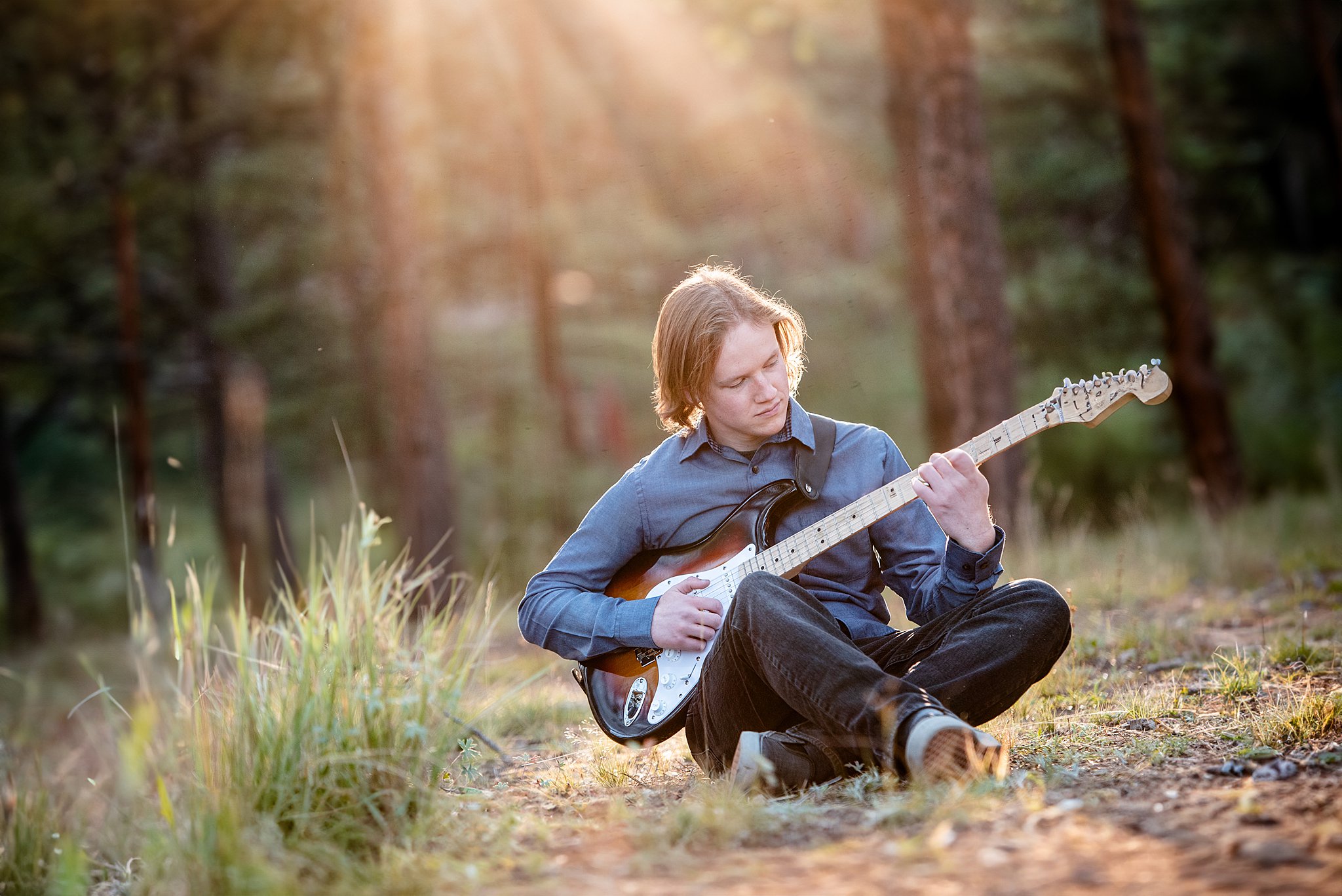 A high school senior plays a guitar while sitting in a forest at sunset