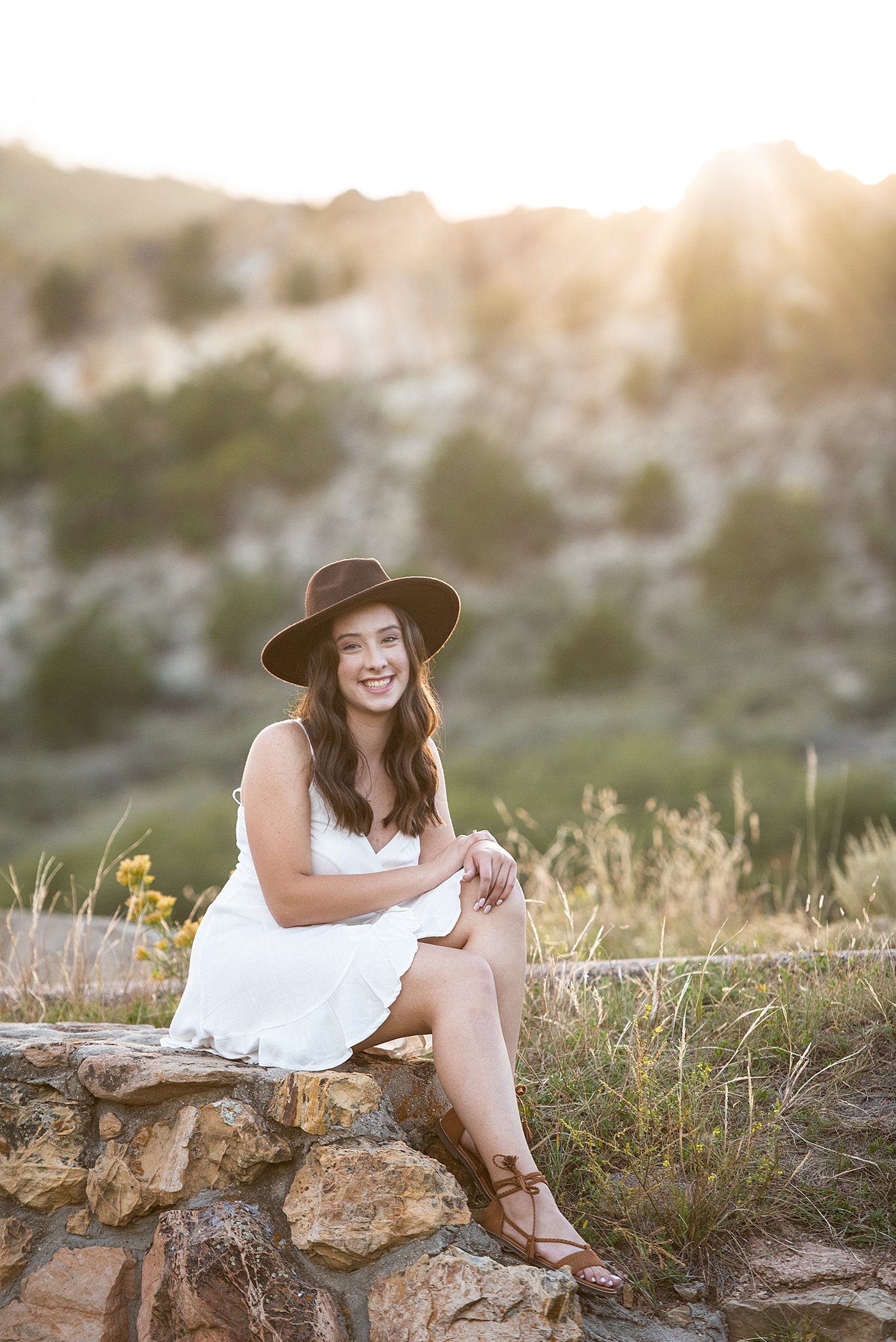 A high school senior in a white dress and brown hat sits on a stone wall in a park at sunset after getting some tutoring colorado springs