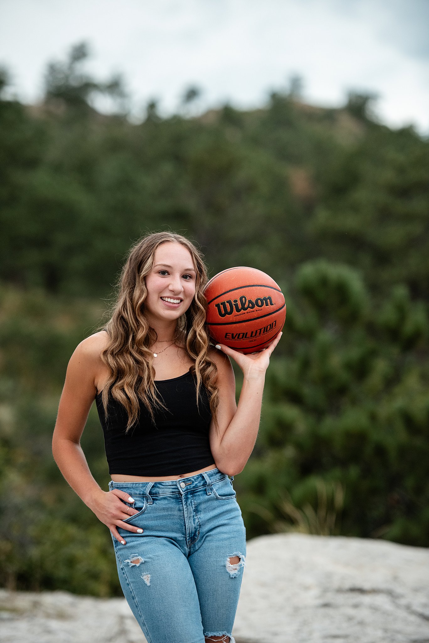 A high school senior in jeans and a black shirt holds a basketball while standing in a rock in a park