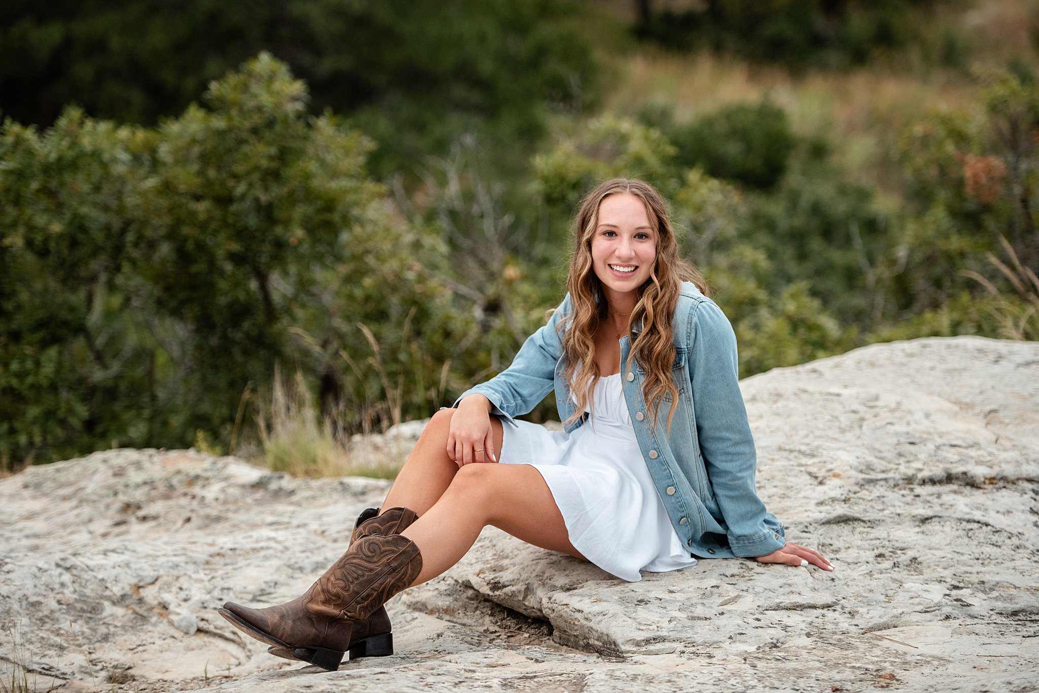 A high school senior in cowboy boots, a white dress and jean jacket sits on a large rock