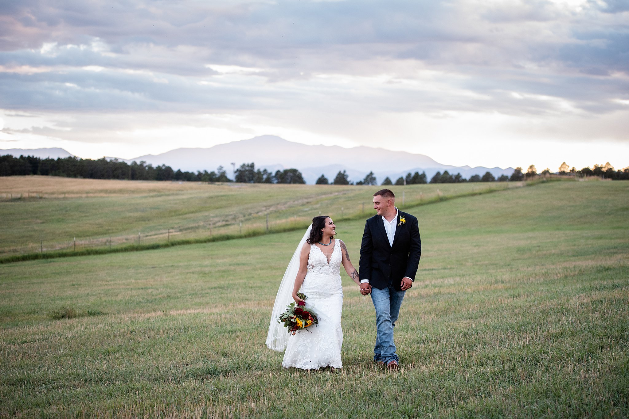 Newlyweds walk hand in hand through a pasture smiling at each other at a younger ranch wedding