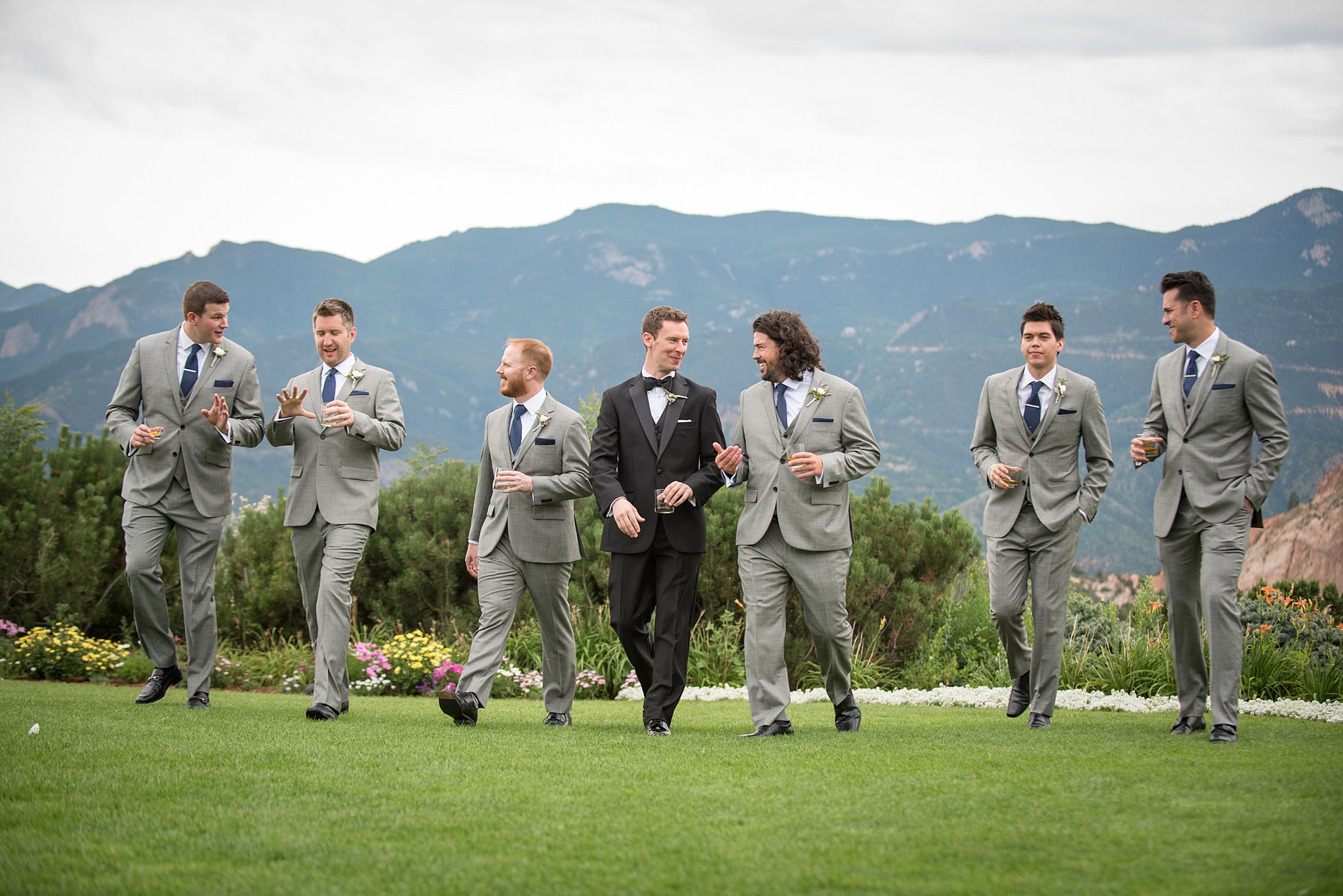 A groom walks in a lawn in a black tux with his groomsmen in grey suits