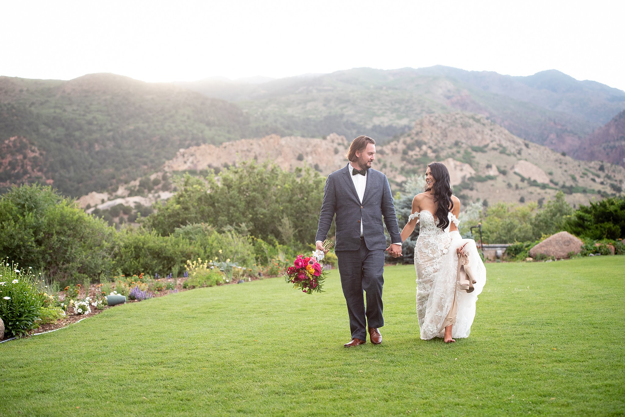 Newlyweds walk in the lawn as the groom holds the colorful bouquet and bride holds her long train and shoes