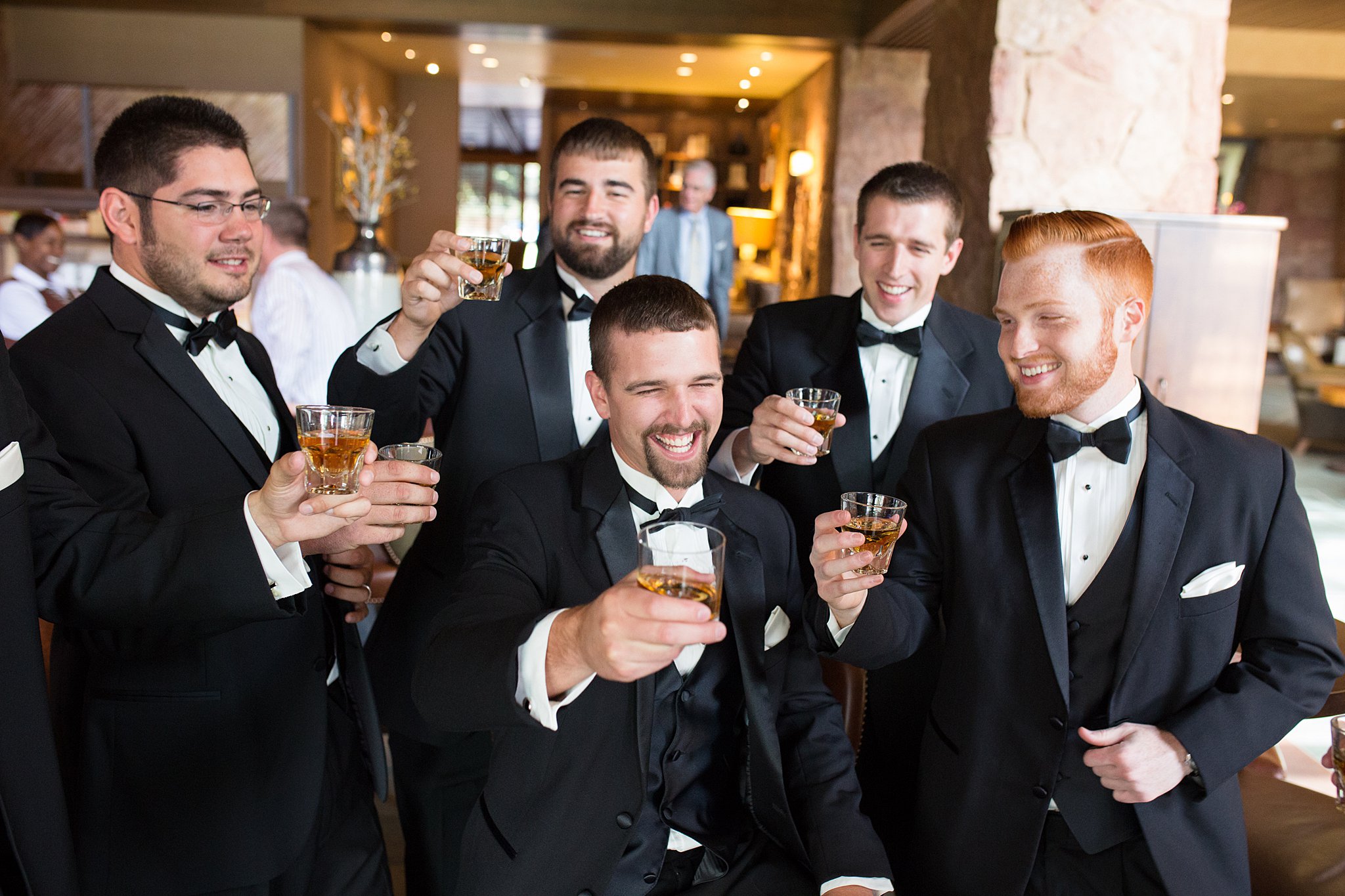 A groom toasts a shot of whiskey with his groomsmen all in matching black tuxedos