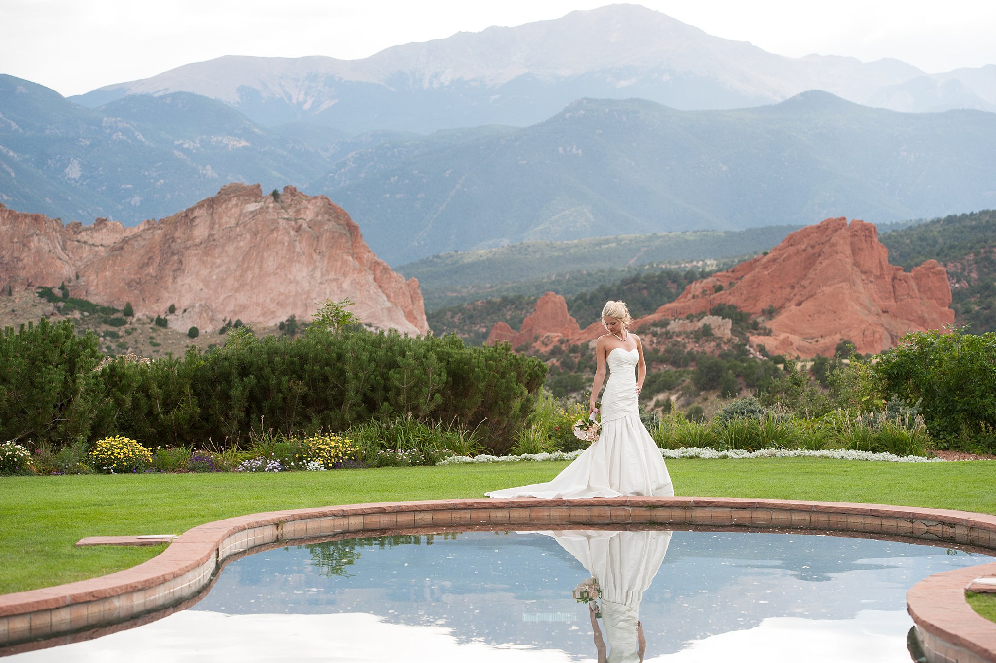 A bride gazes down at her train and bouquet as she walks along the pool at her Garden Of The Gods Club Wedding