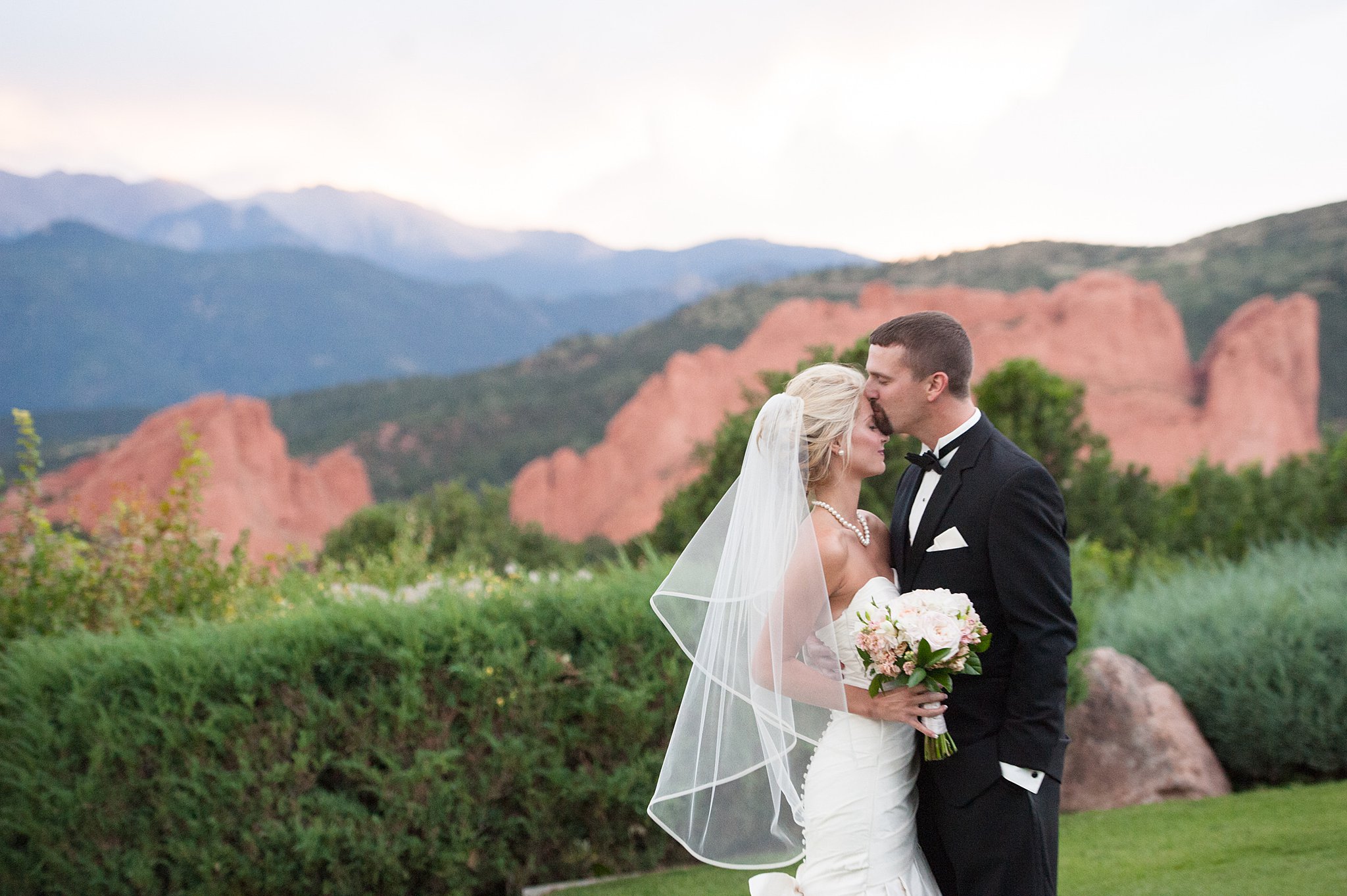 A groom in a black tux kisses his bride's forehead in the lawn at their Garden Of The Gods Club Wedding