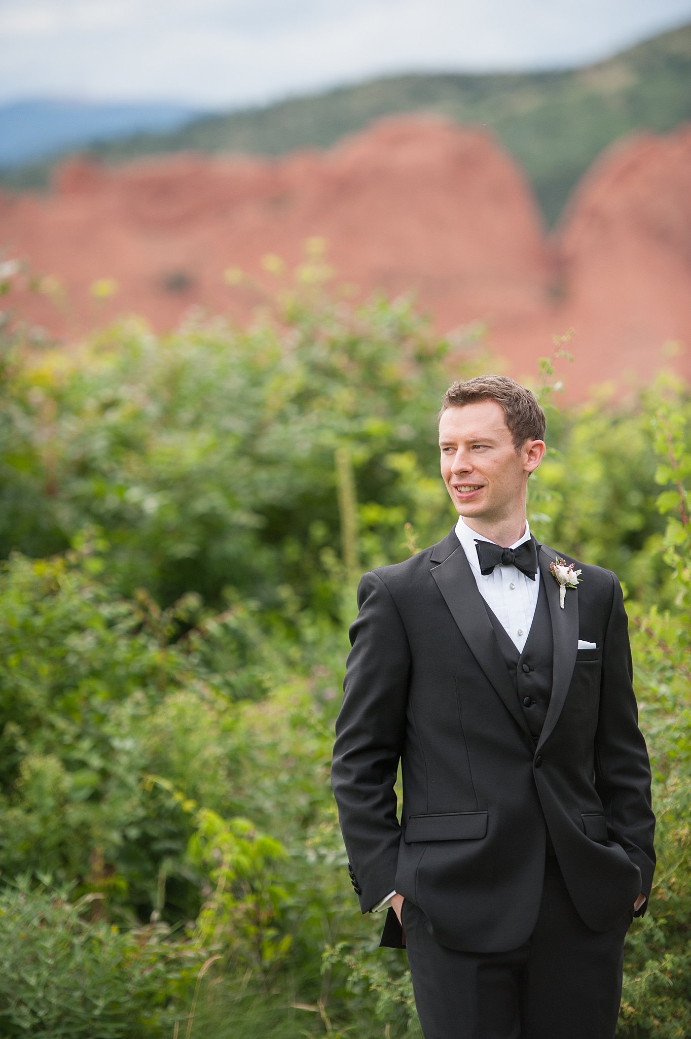 A groom stands in the garden with hands in pockets smiling over his shoulder