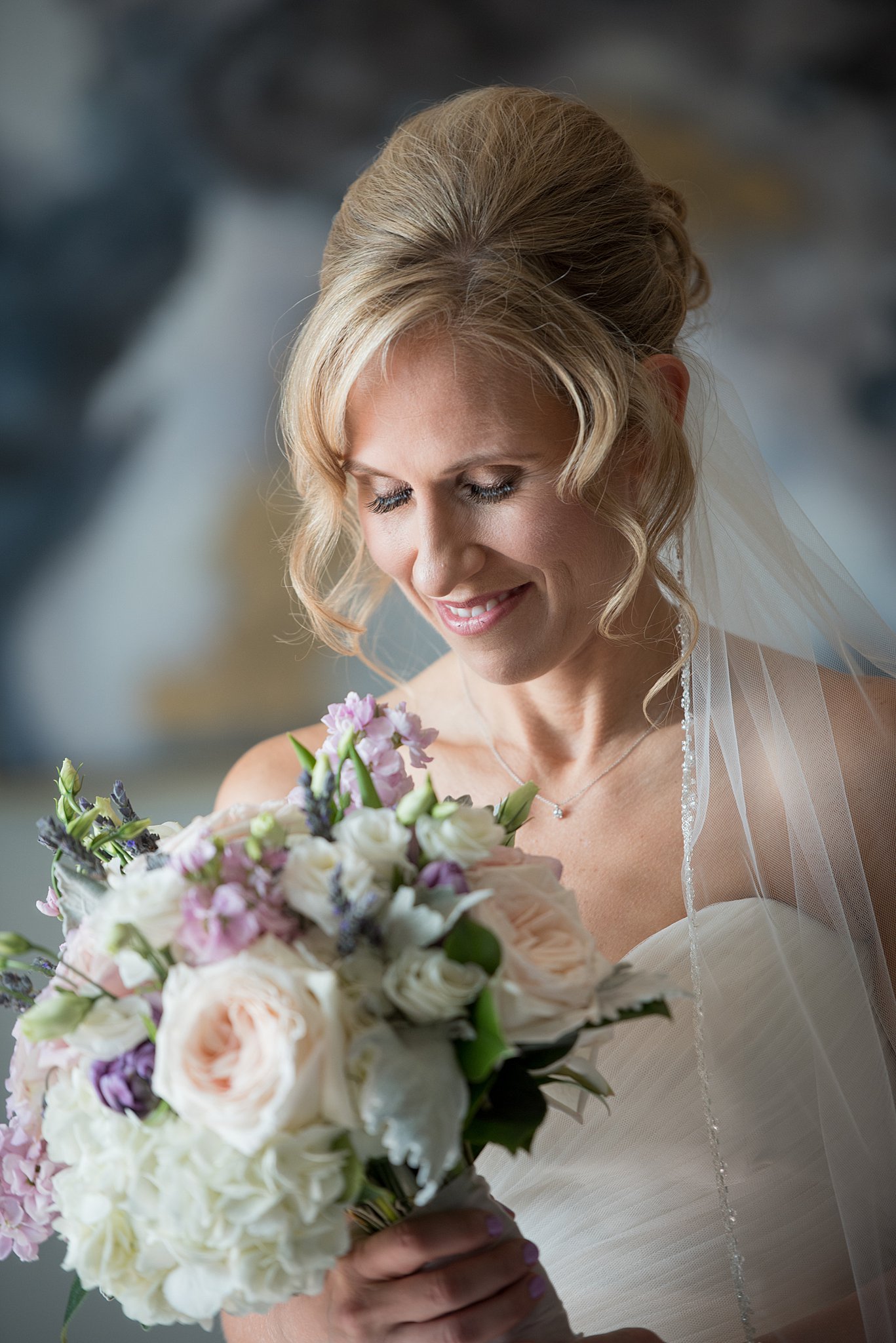 A bride smiles down at her white, pink and purple bouquet