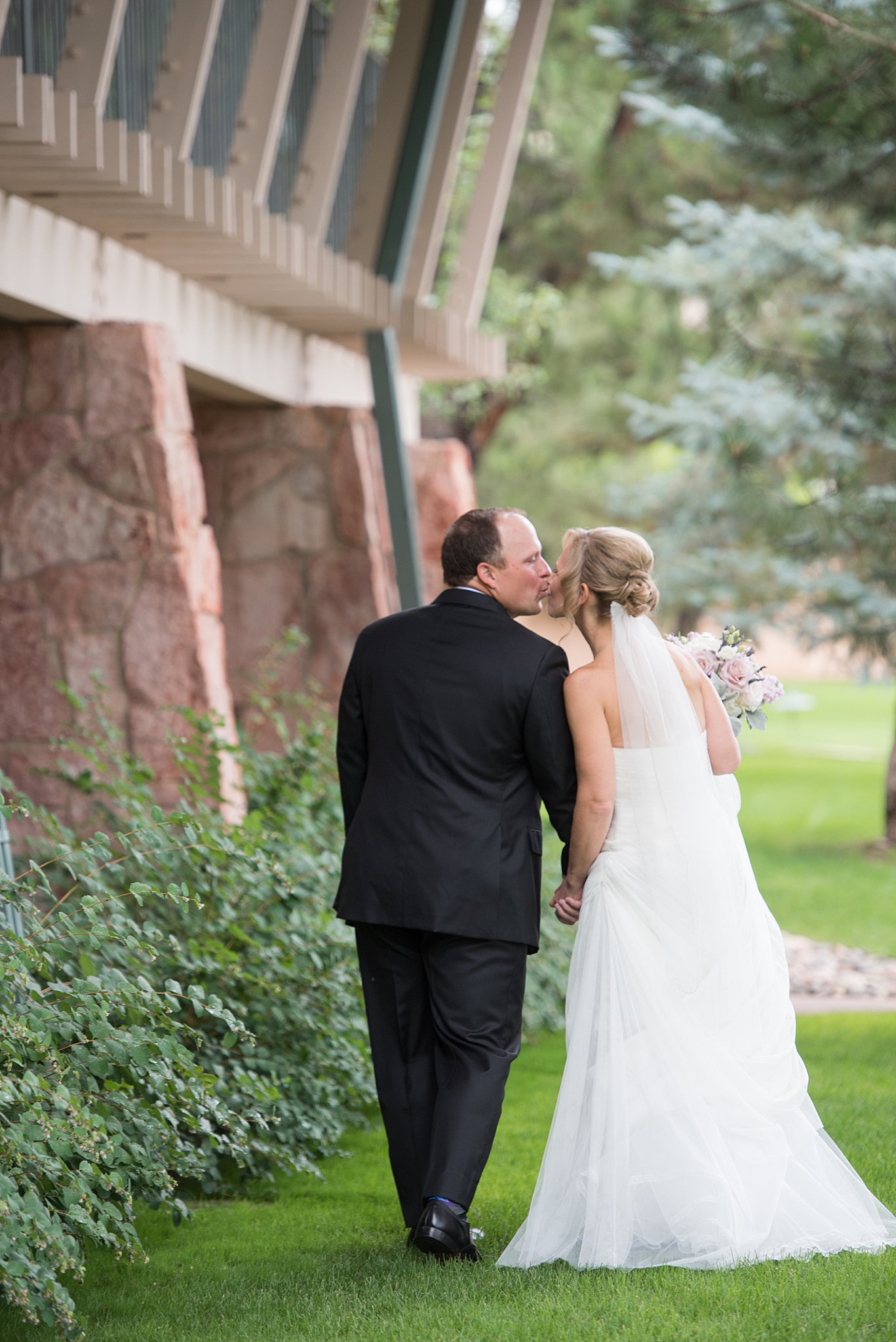 A bride and groom kiss while walking in the lawn and holding hands