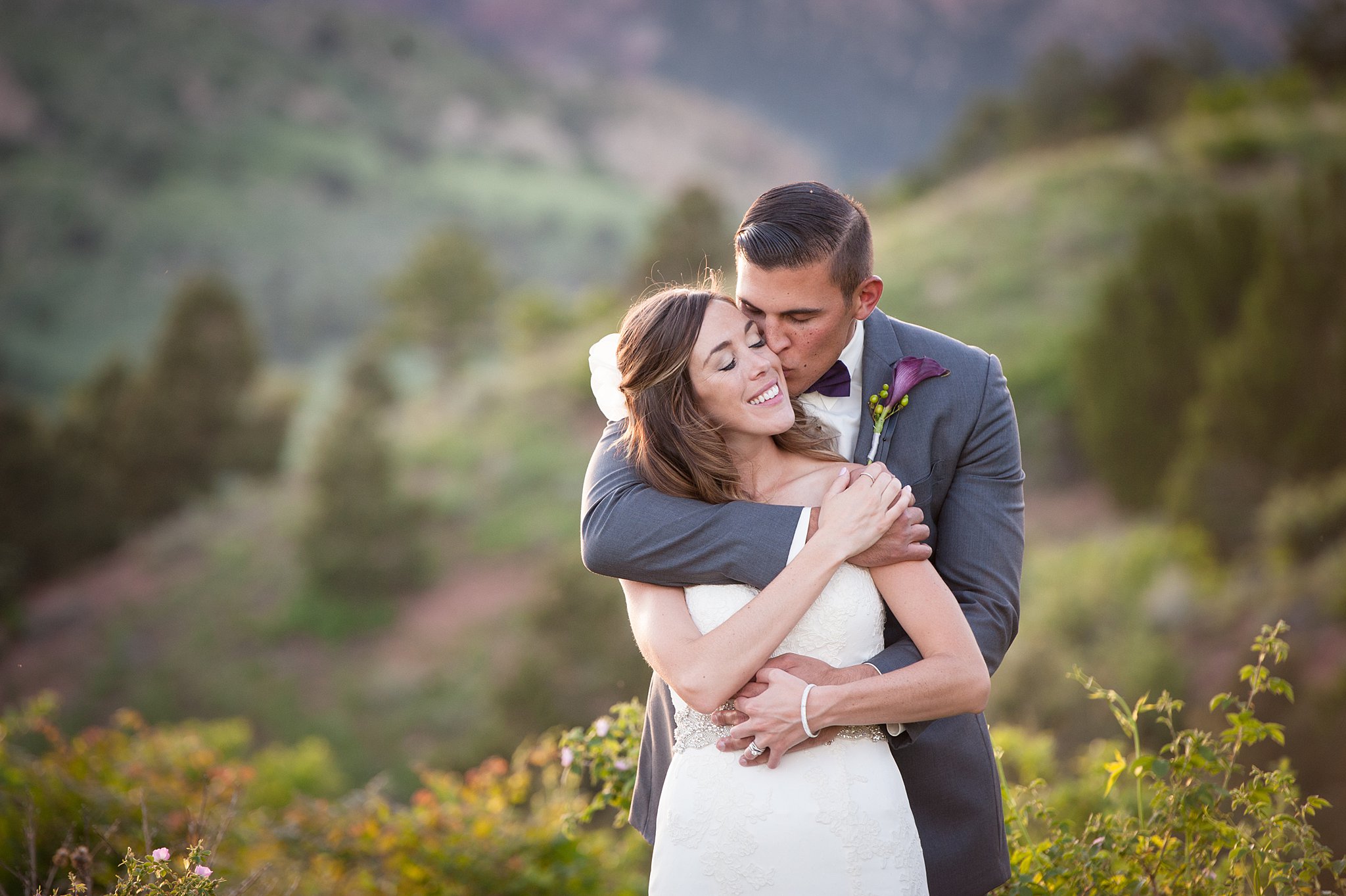 A groom hugs his bride from behind and kisses her cheek at sunset