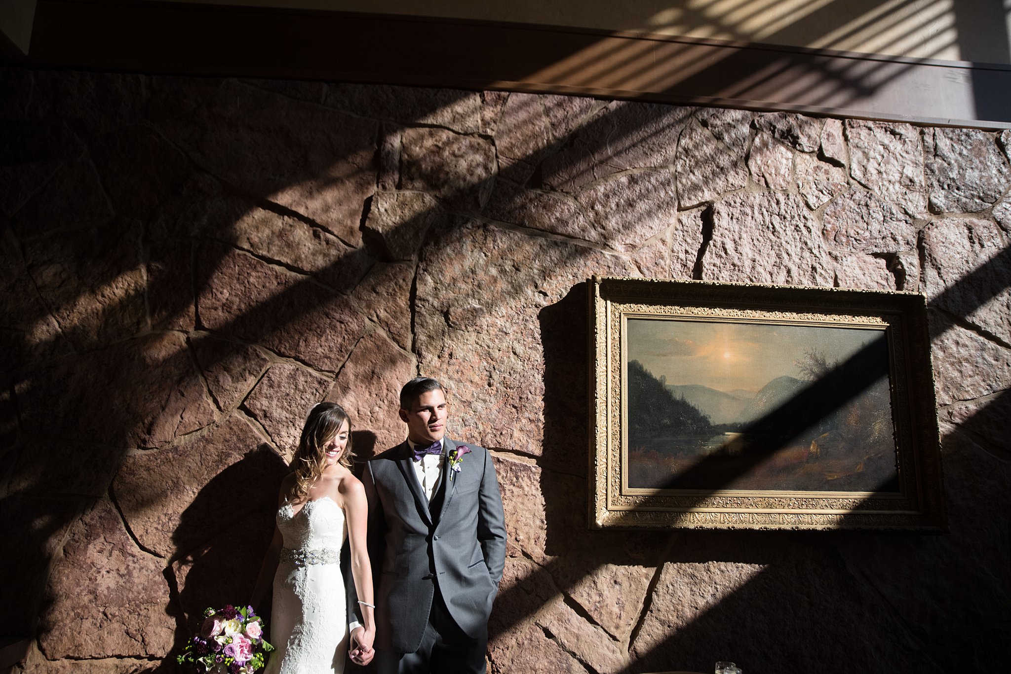 A bride and groom lean on a stone wall with dramatic lighting holding hands and smiling