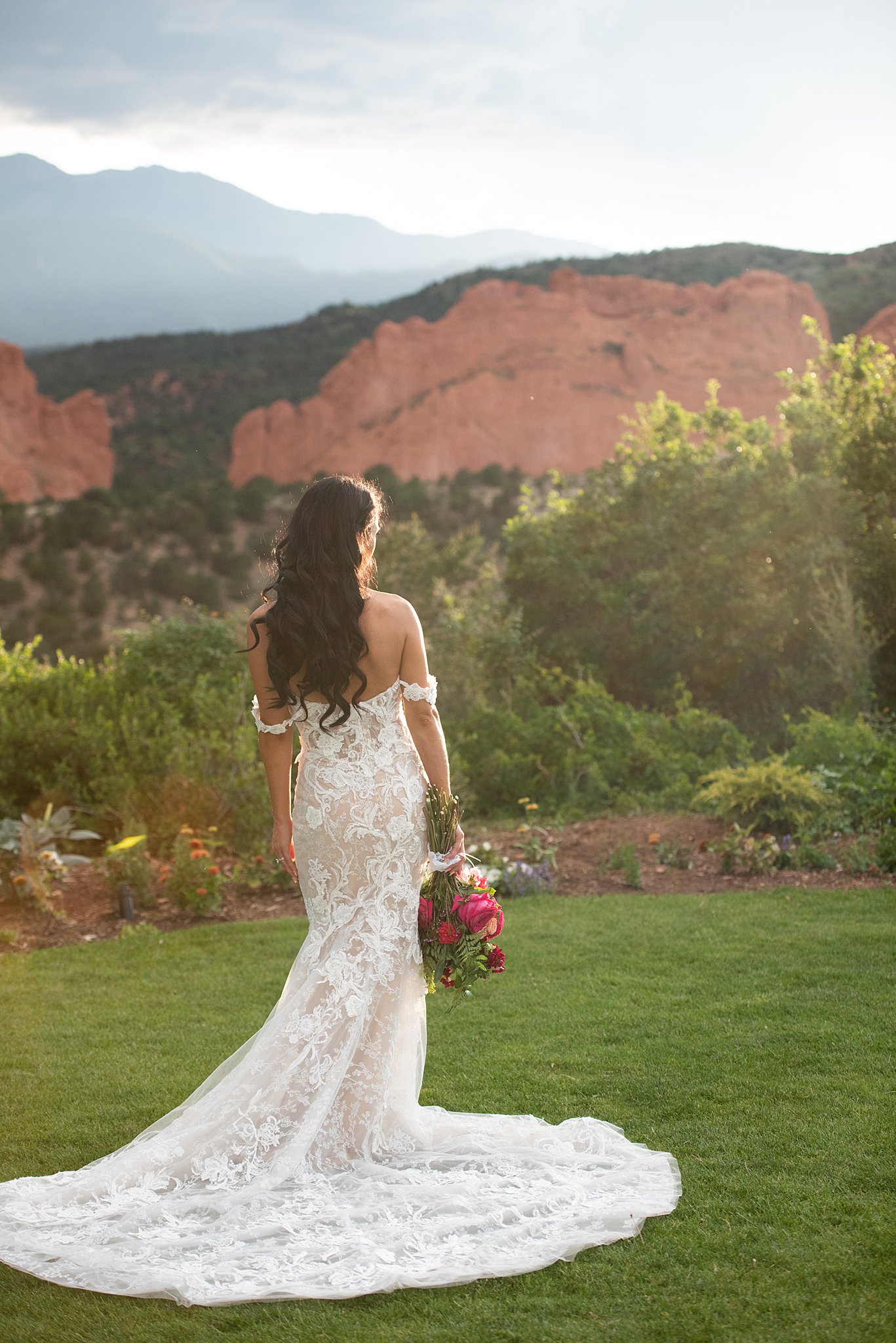 A bride stands in her lace dress with long train and red bouquet at her Garden Of The Gods Club Wedding at sunset