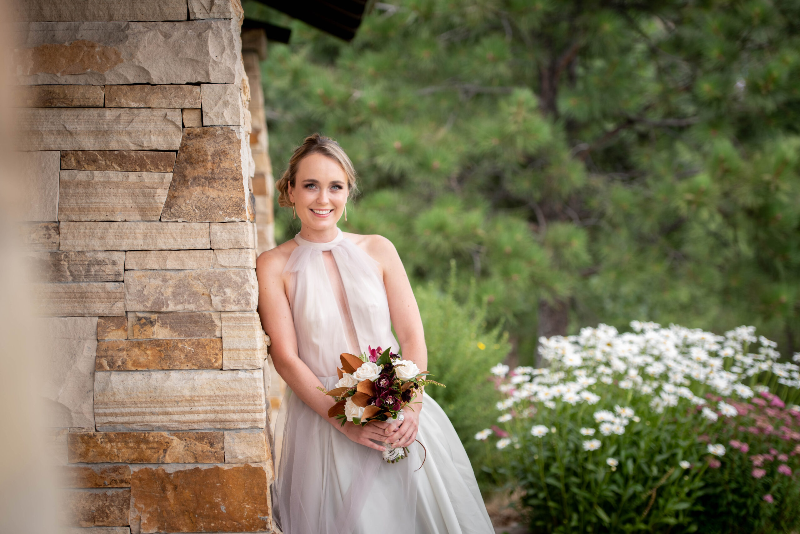 A smiling bride leans on a stone wall in a garden at the Spruce Mountain Ranch wedding venue holding her bouquet