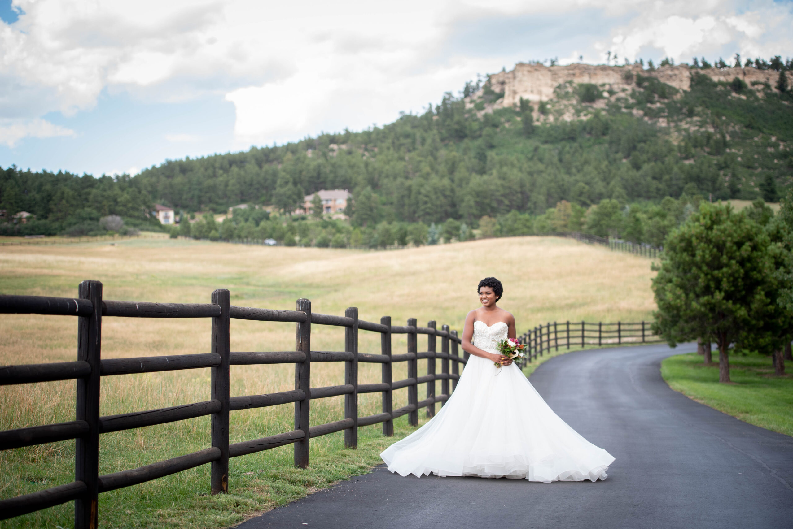 A happy bride walks on a paved sidewalk with her bouquet along a pasture at the Spruce Mountain Ranch wedding venue