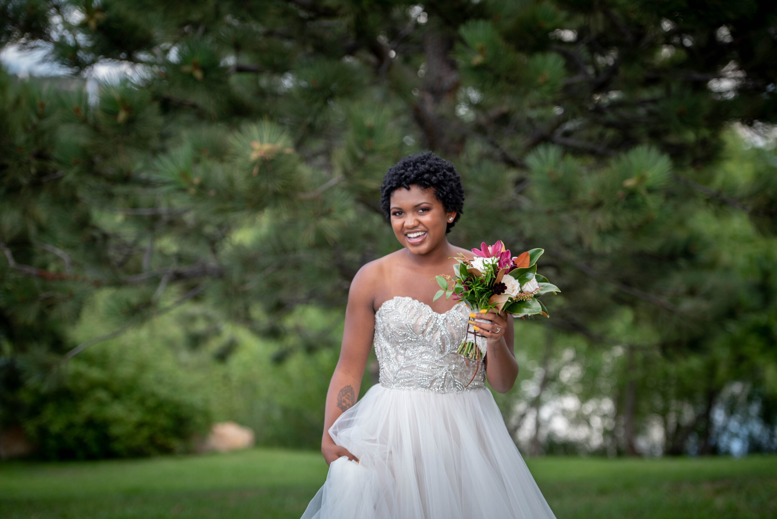 A happy bride plays in her dress under some trees with a colorful bouquet