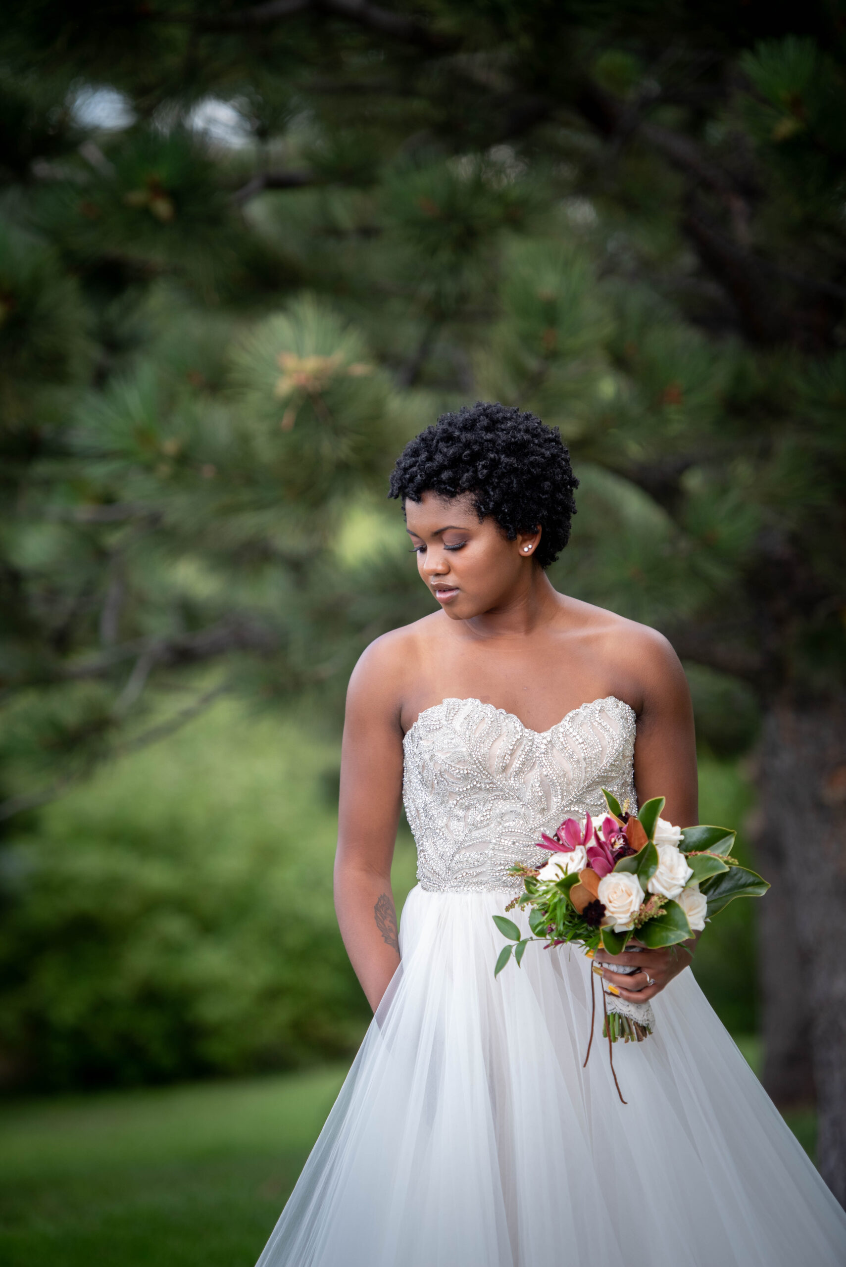 A bride smiles down her shoulder while holding a pink and white bouquet under a tree