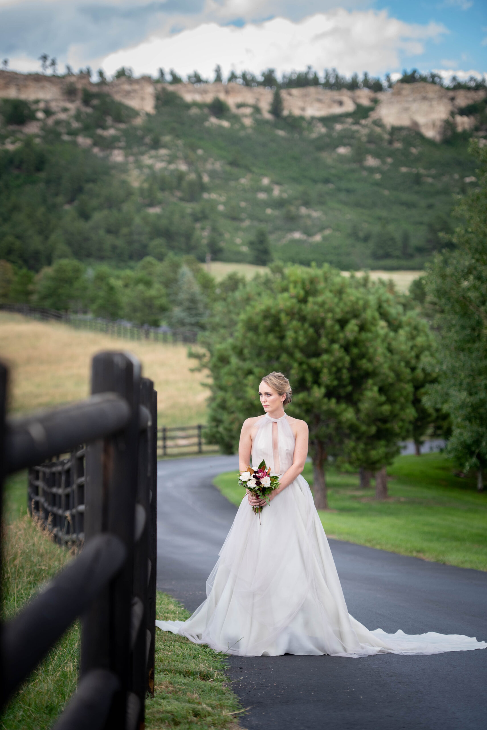 A bride walks a sidewalk along a wooden pasture fence holding her bouquet