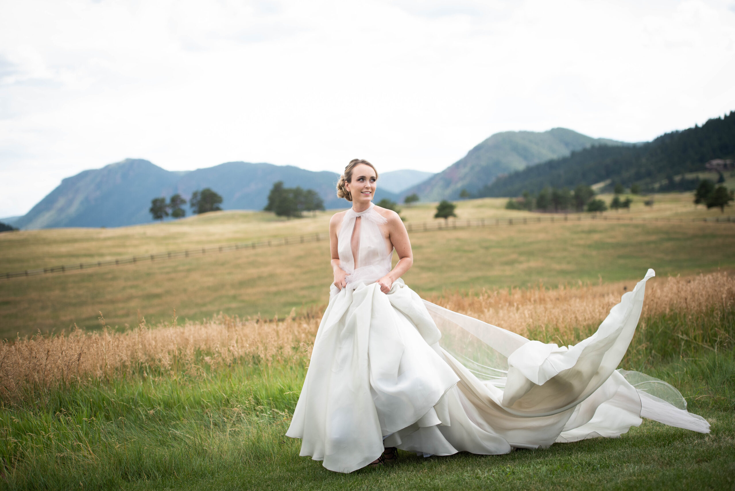 A bride's train blows in the wind at the Spruce Mountain Ranch wedding venue as she smiles in a pasture at sunset