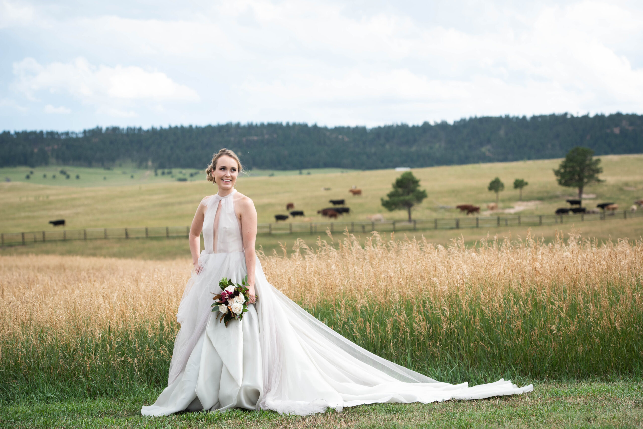 A bride smiles with a hand on her hip on the edge of tall golden grass in a pasture