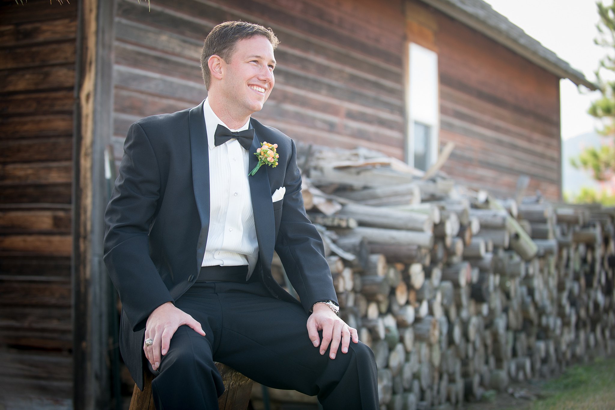 A happy groom at the Spruce Mountain Ranch wedding venue in a black suit sits by a cord of firewood smiling