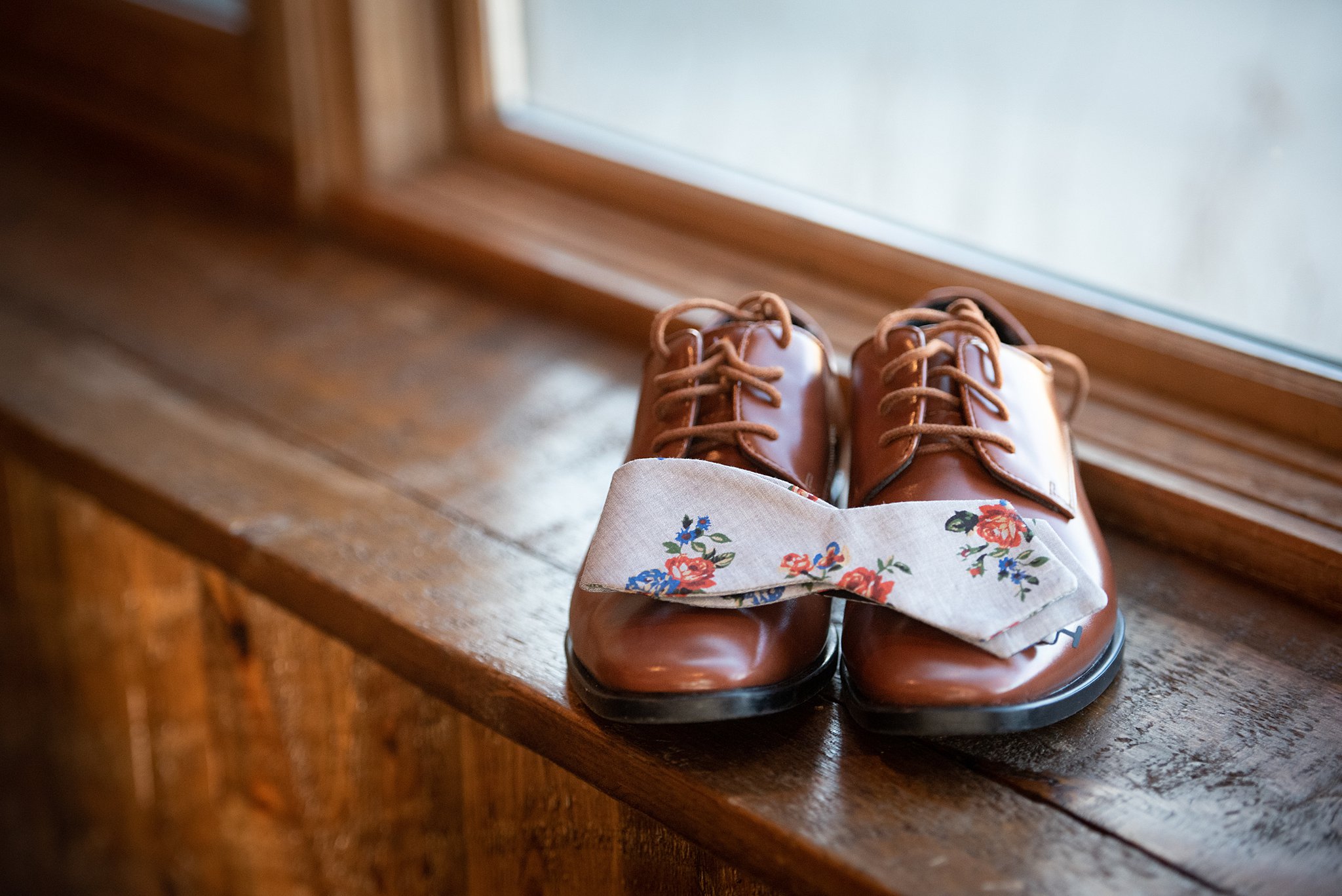 Details of a groom's brown shoes and floral bowtie in a window sill