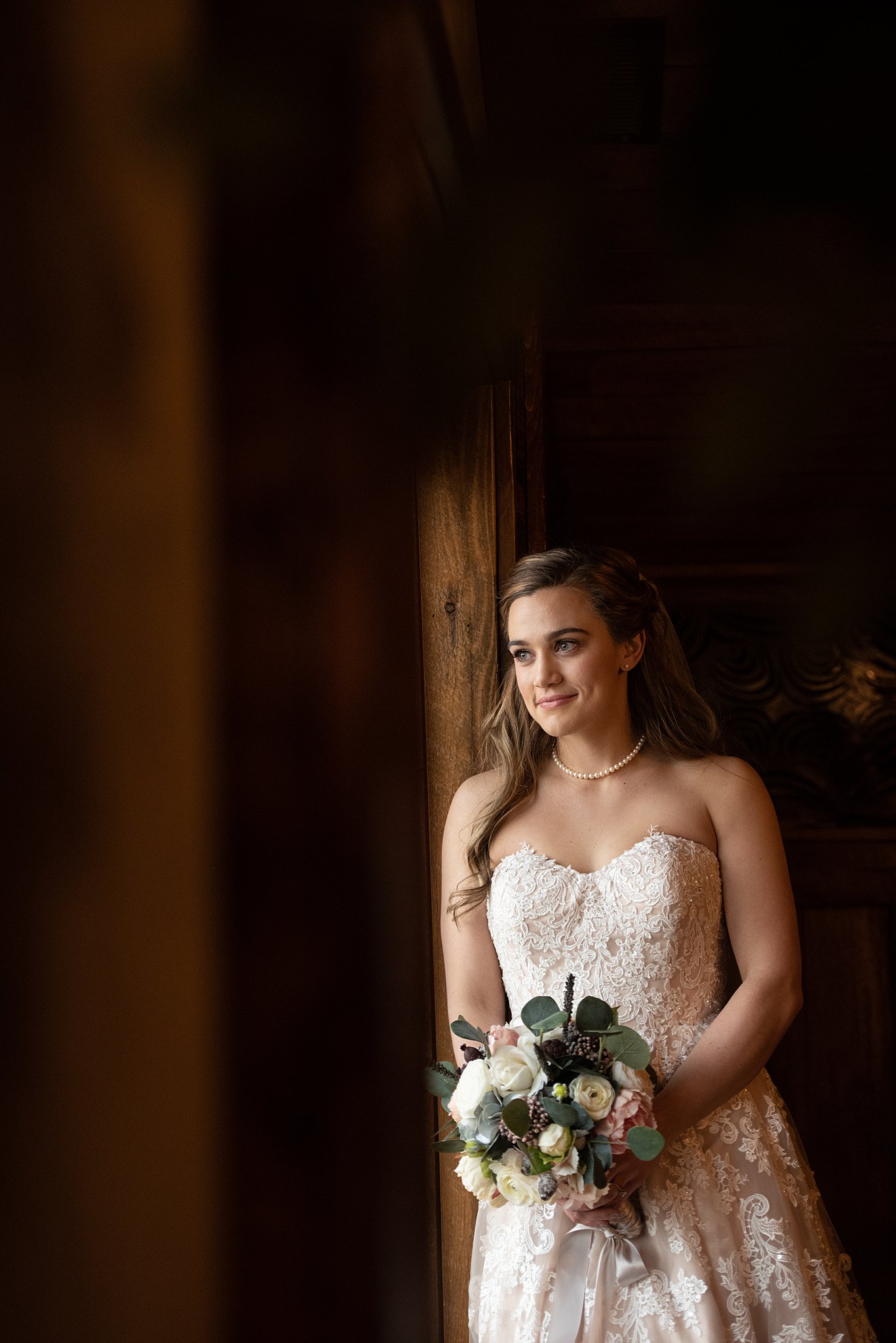 A bride at a Spruce Mountain Ranch wedding leans in a window frame smiling while holding her bouquet