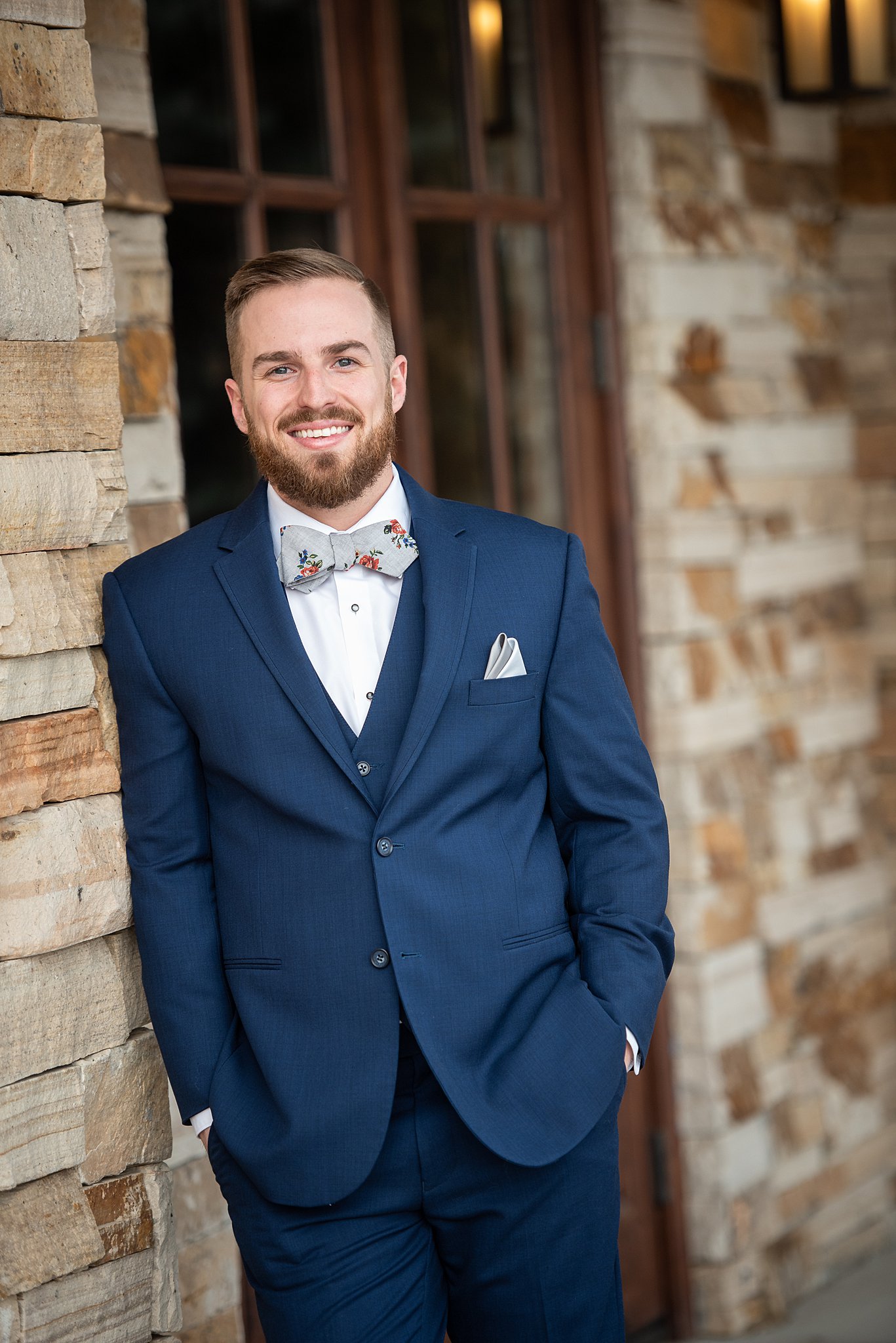 A smiling groom in a blue suit and floral bowtie leans on a stone wall