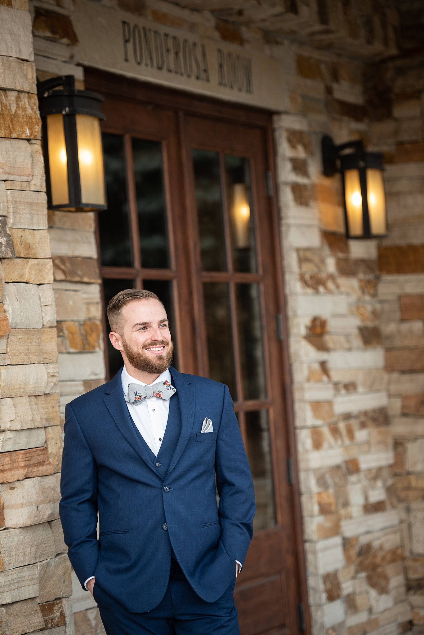 A groom leans on a stone wall by entrance door in a blue suit and floral bowtie