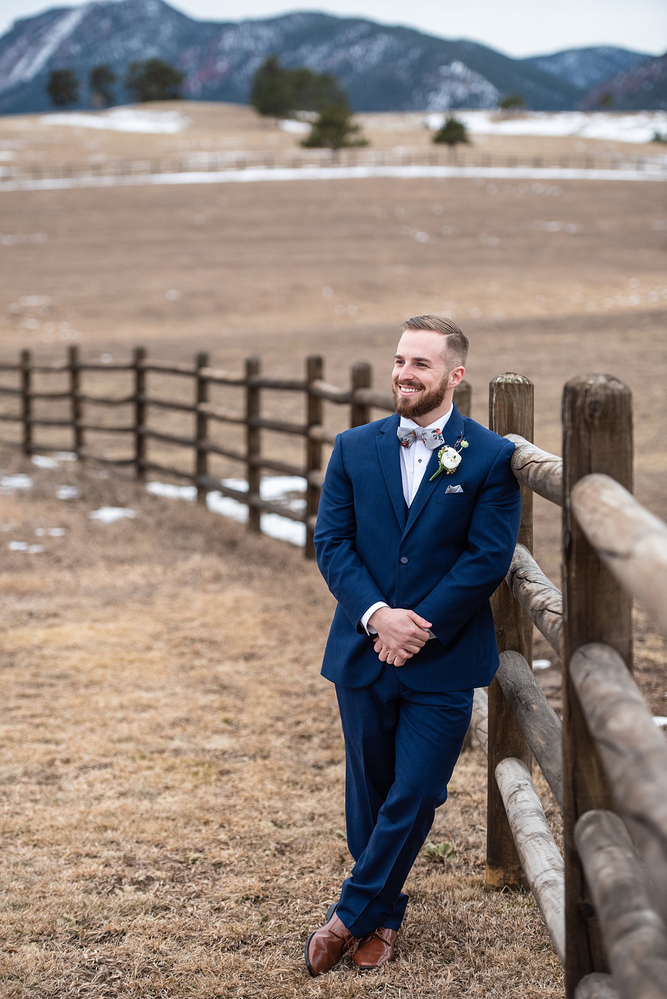 A groom leans on a pasture wooden beam fence in a blue suit at his Spruce Mountain Ranch wedding