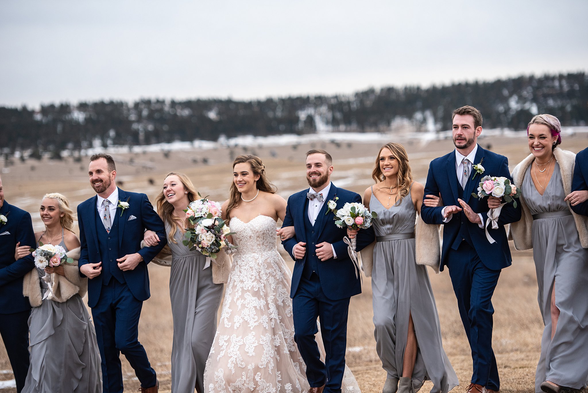 A bride and groom lock arms with their laughing bridal party while walking in a pasture at their Spruce Mountain Ranch wedding