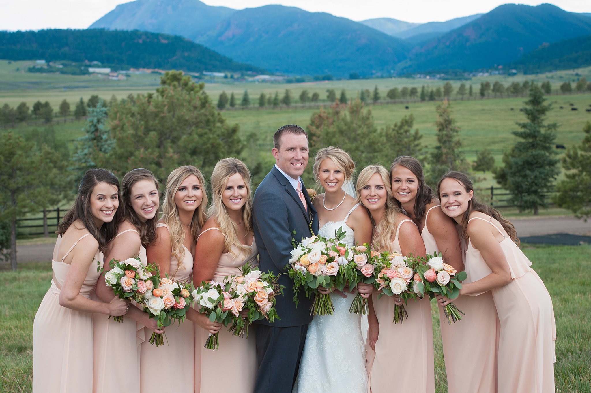 Newlyweds stand smiling with their bridesmaids in pink dresses in a meadow with mountains backdrop at Spruce Mountain Ranch wedding venue