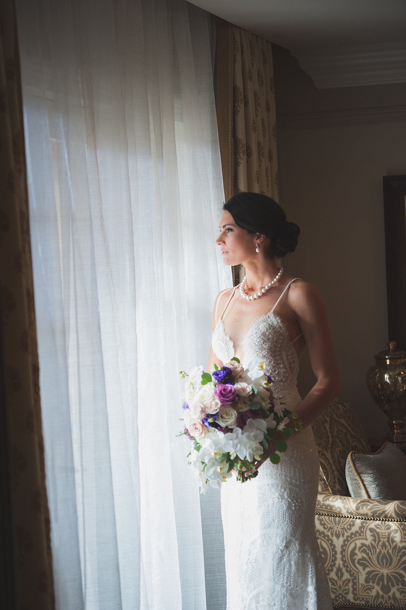 A bride gazes out a window while holding her purple and white bouquet